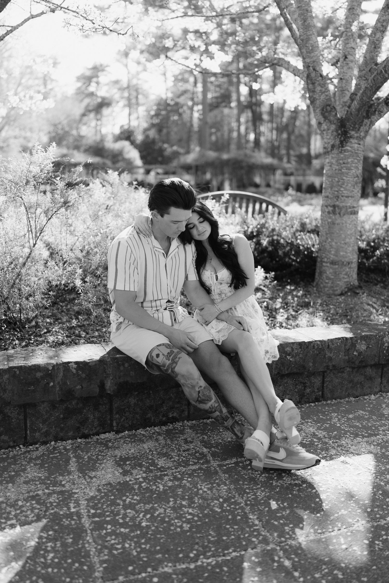 Black and white photo of a couple sitting close together on a stone wall in a lush garden setting.