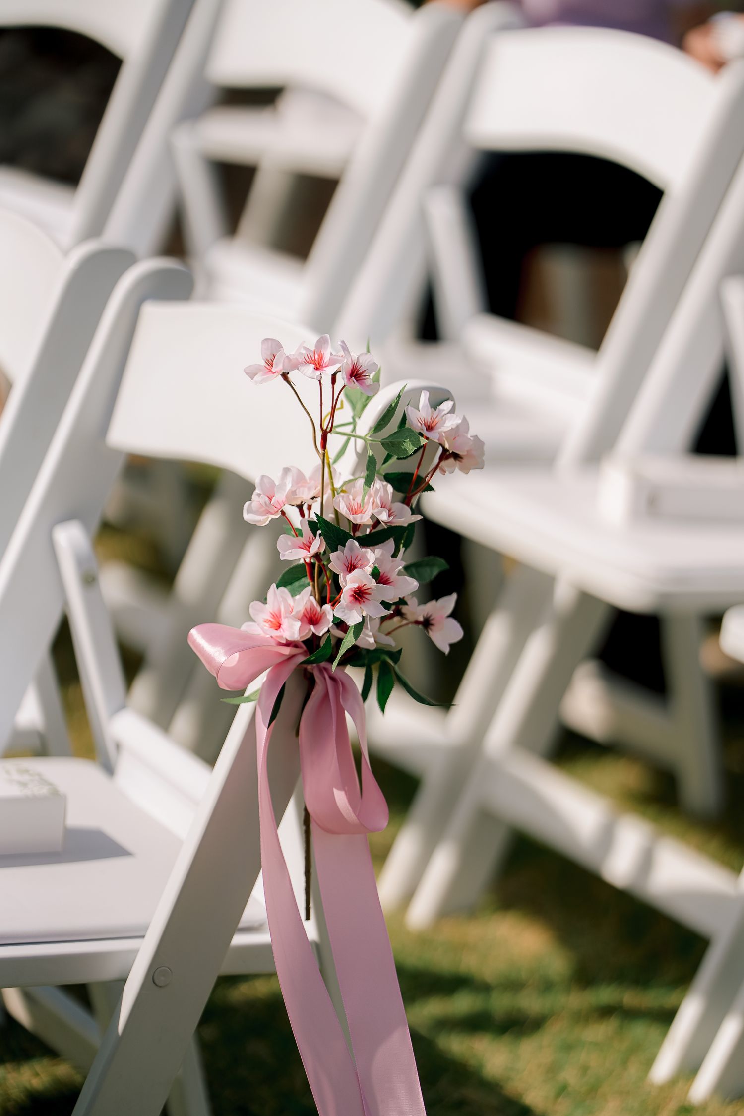 Pink floral bouquet with ribbon tied to white folding chair at outdoor wedding ceremony aisle decoration.
