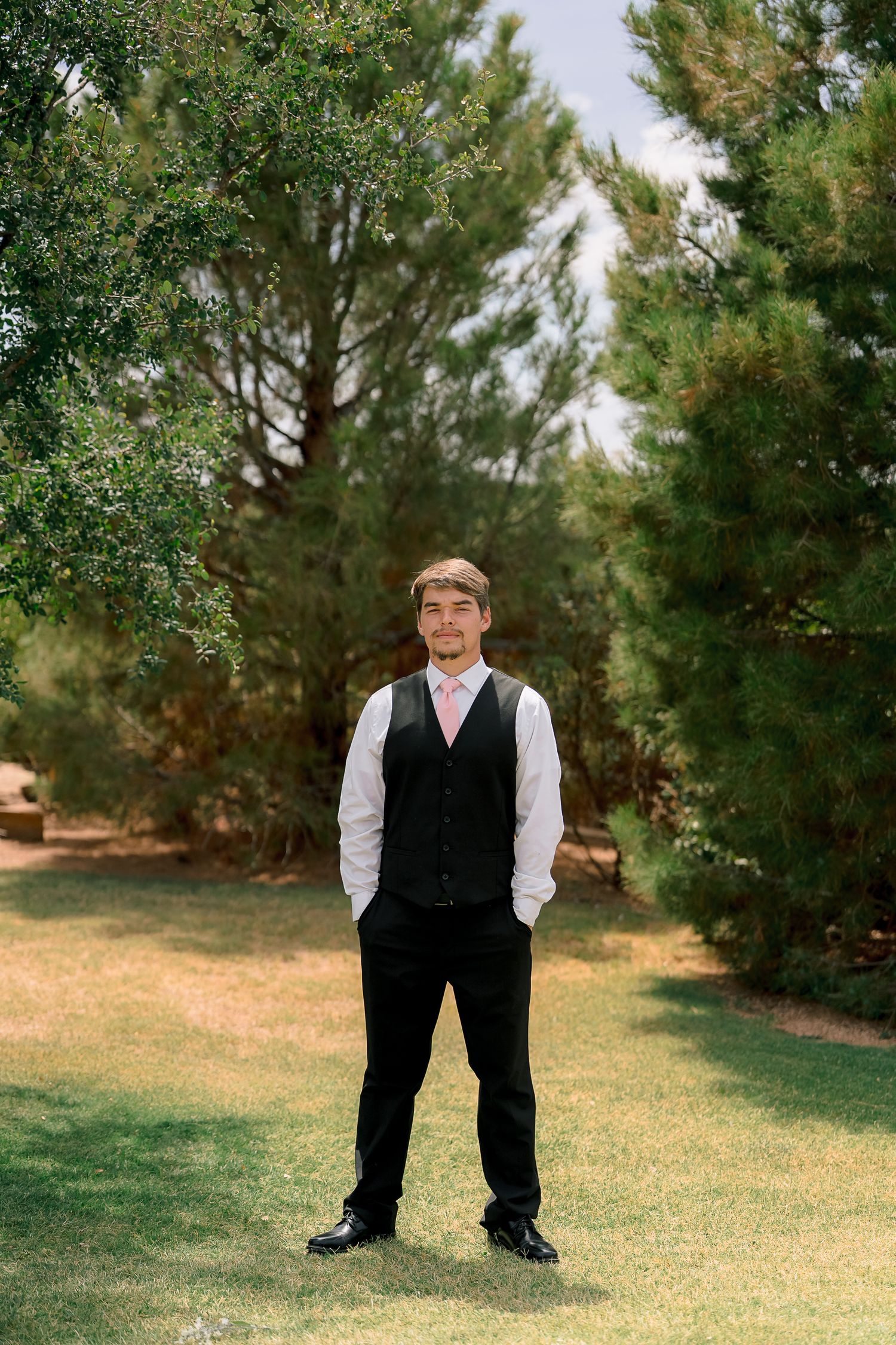 Groom in black vest and pink tie stands on green lawn surrounded by tall pine trees on wedding day.