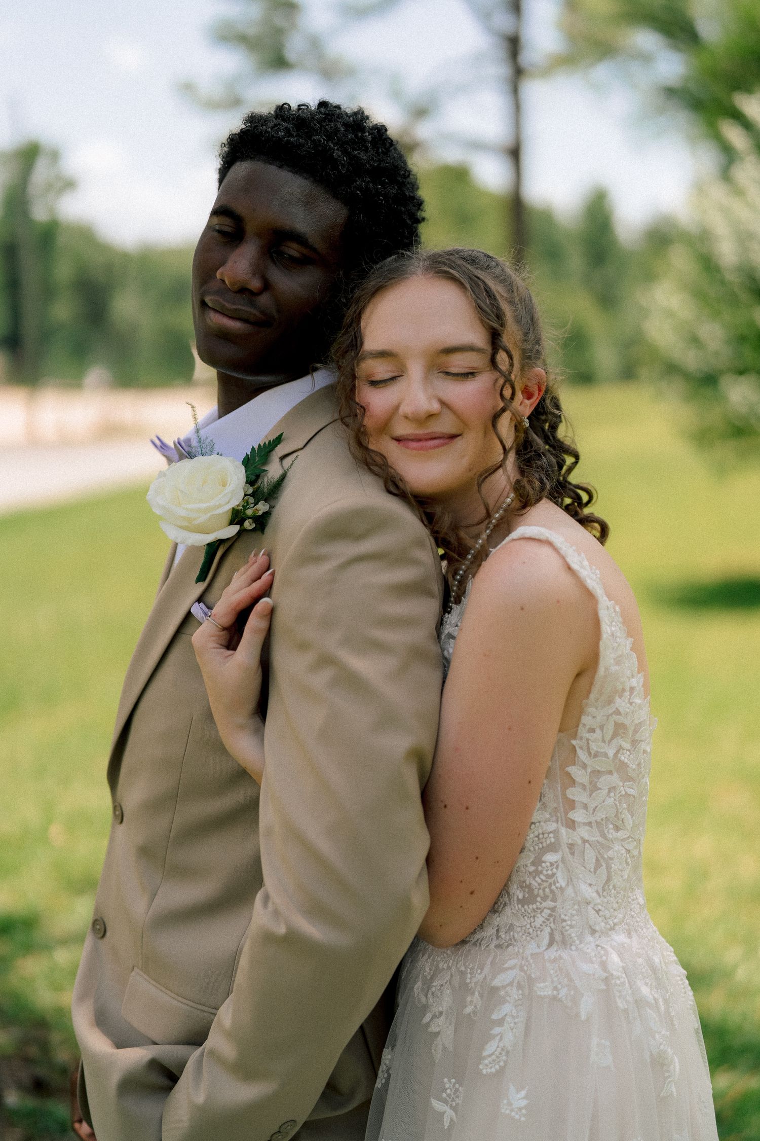 Bride in lace wedding gown rests her head on groom's shoulder outdoors, groom wearing tan suit with white rose boutonniere.