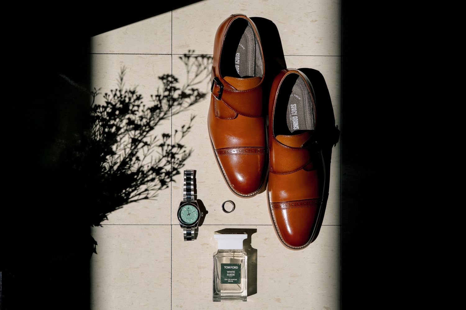Brown leather monk strap dress shoes displayed with a watch, ring, and cologne on a tiled surface in natural light.