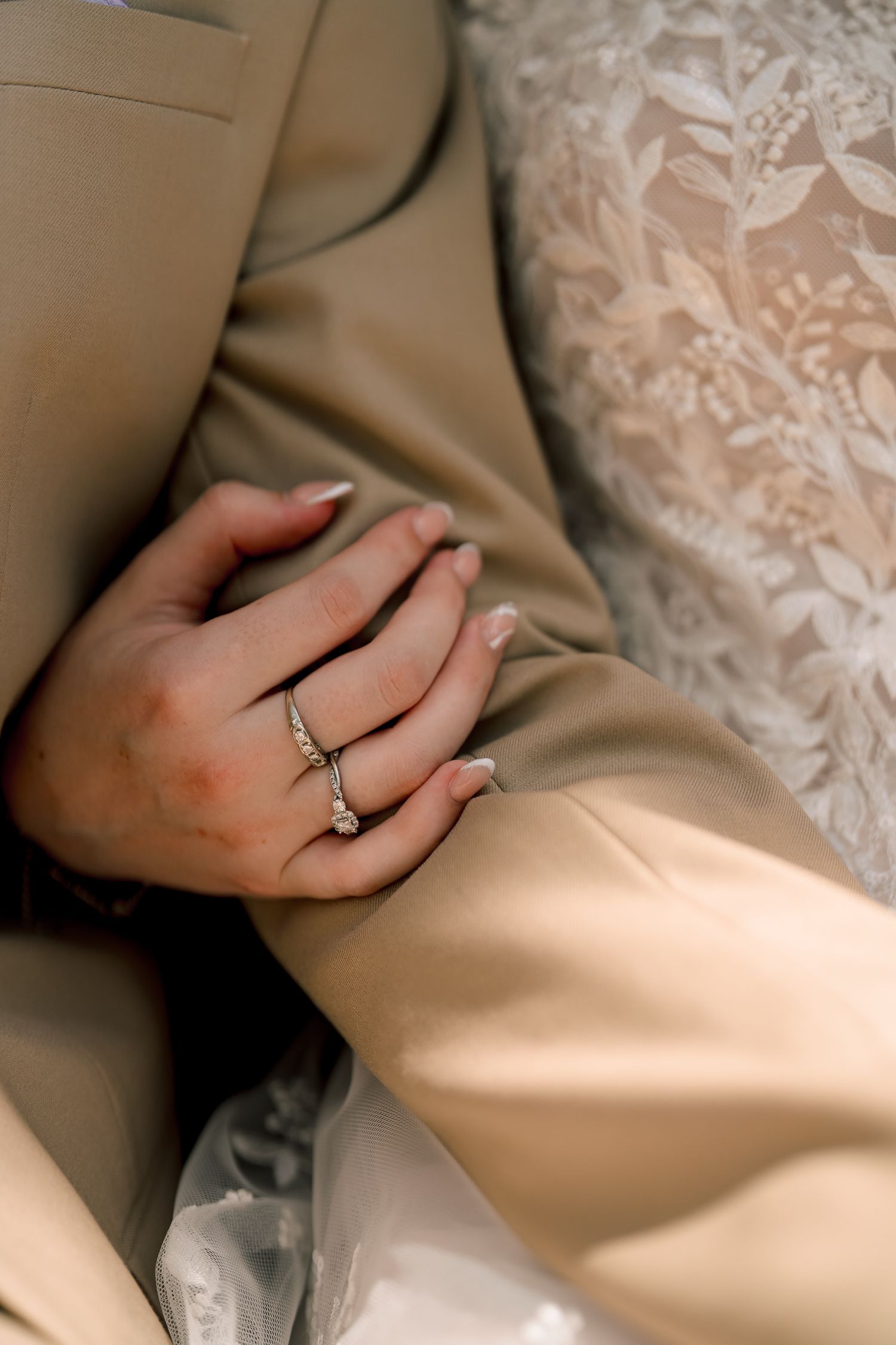 Close-up of bride's hand with diamond engagement ring resting on groom's tan suit jacket.