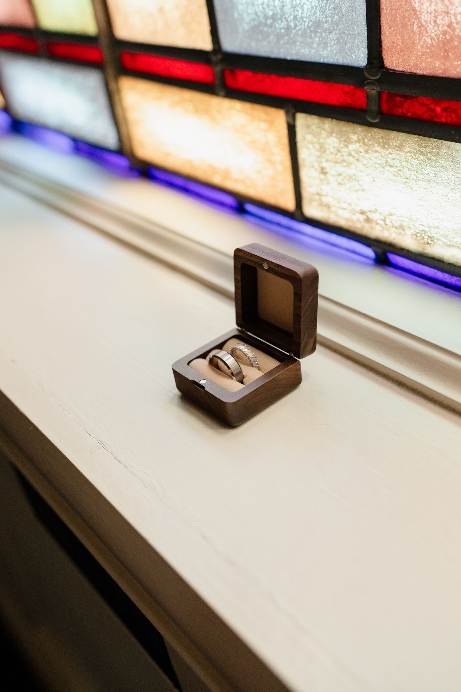 Wedding rings displayed in a small wooden box against a stained glass window background.