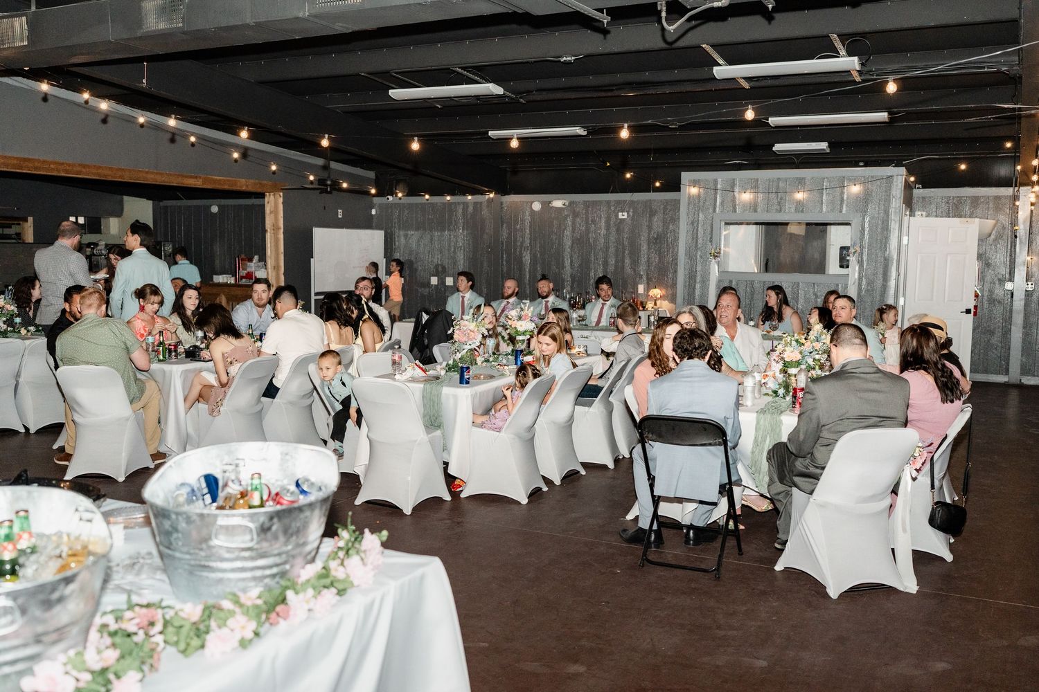 Wedding reception in a modern venue with white chairs, string lights, and floral centerpieces on round tables.