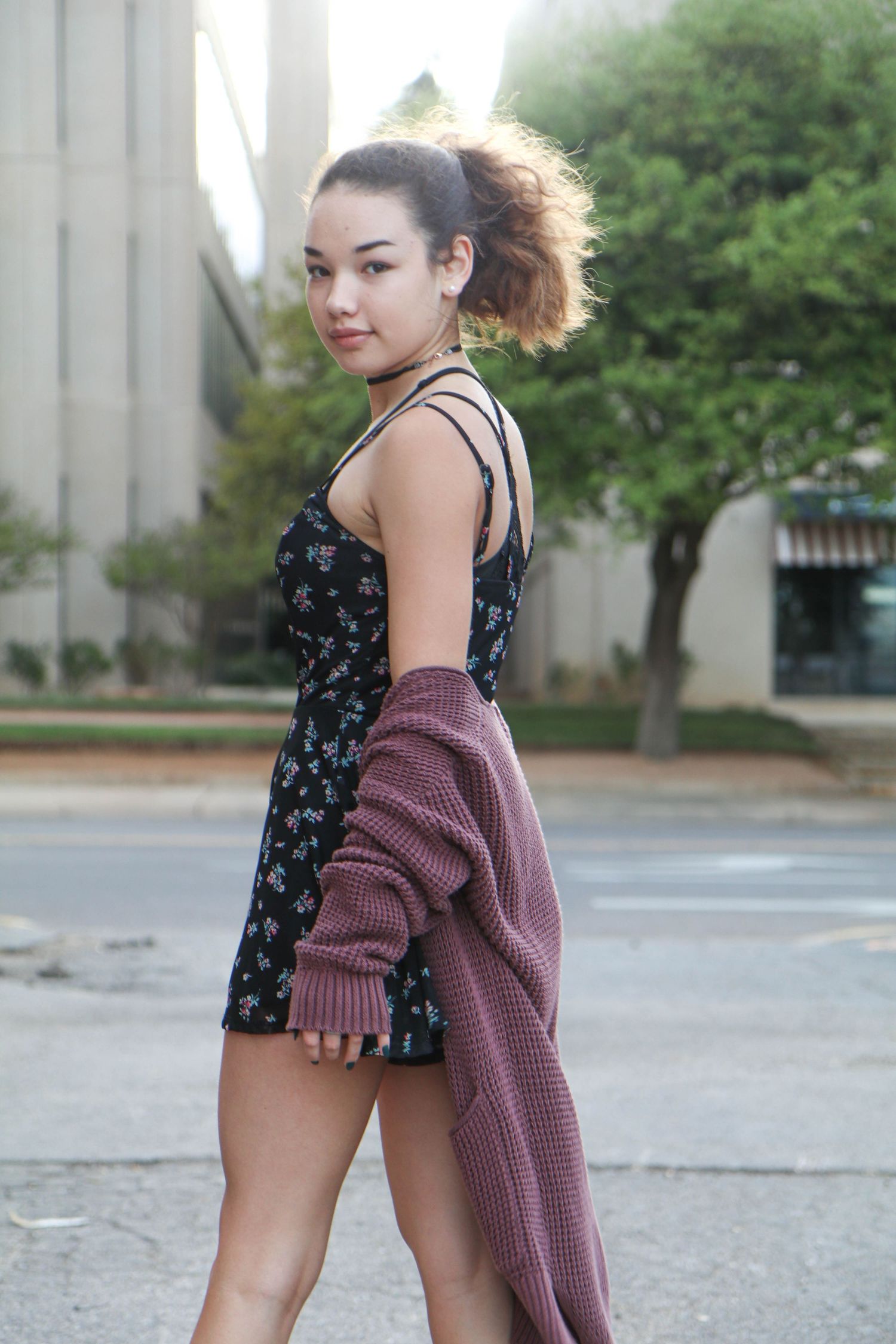 Person in floral romper and sweater posing outdoors near building.