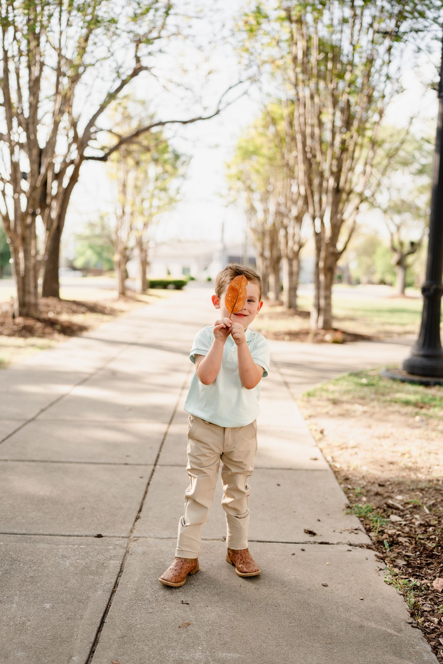 Child in light colored outfit stands on tree-lined sidewalk with hands near face.