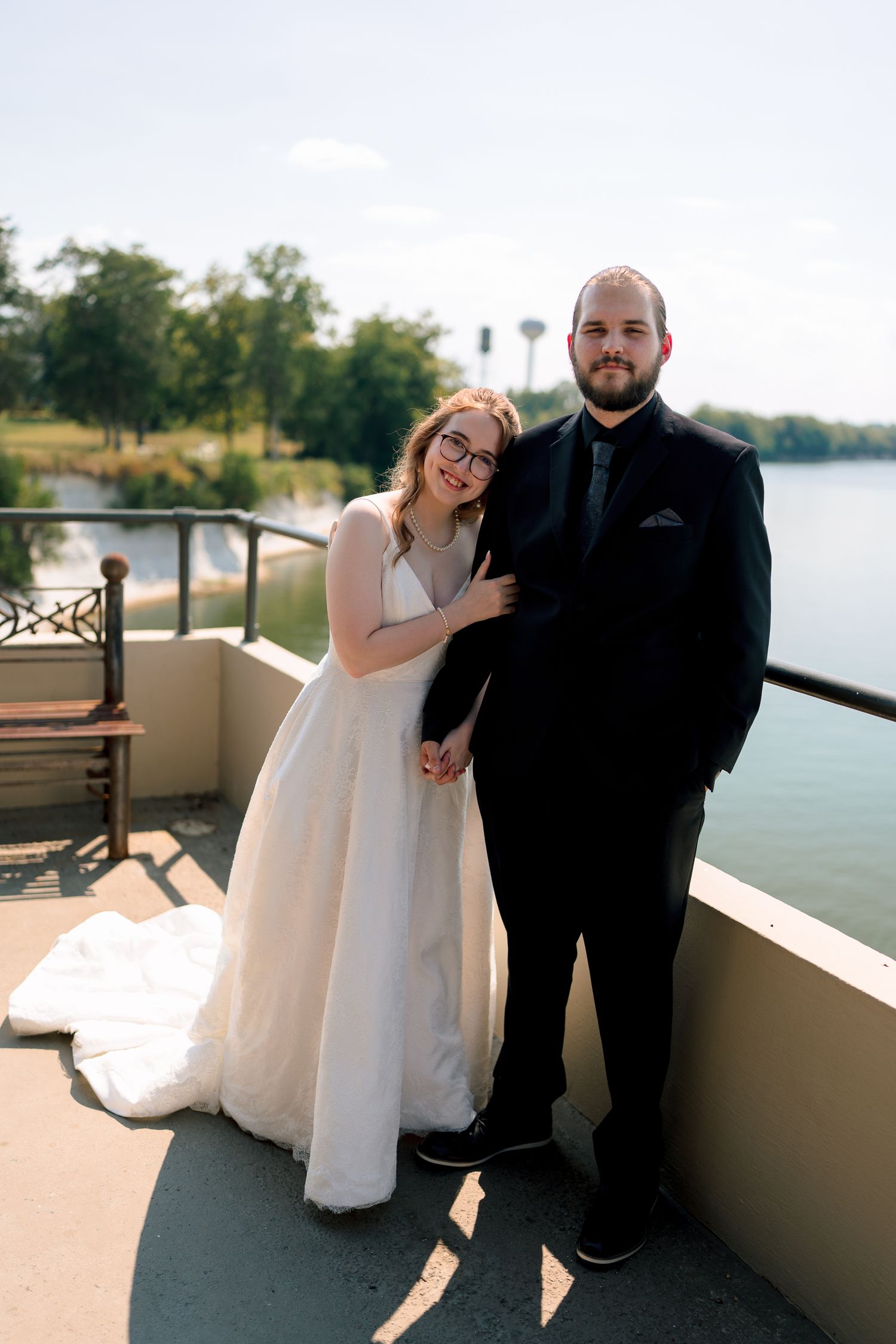 Bride and groom pose on outdoor balcony overlooking a lake, bride in white gown and groom in black suit.