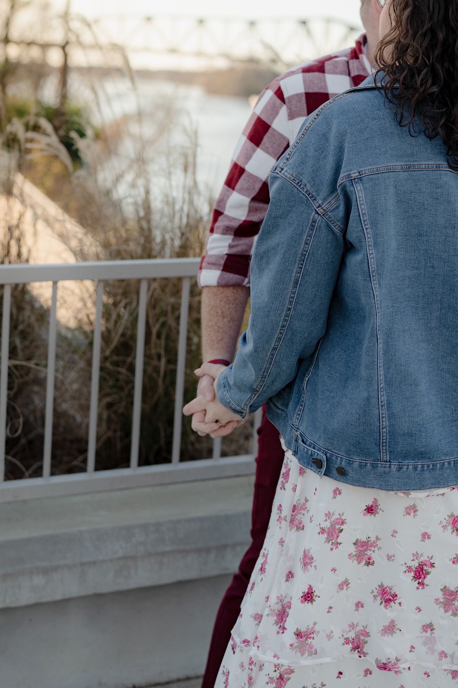 Close-up view of denim jacket and red plaid shirt against railing overlooking river at sunset.