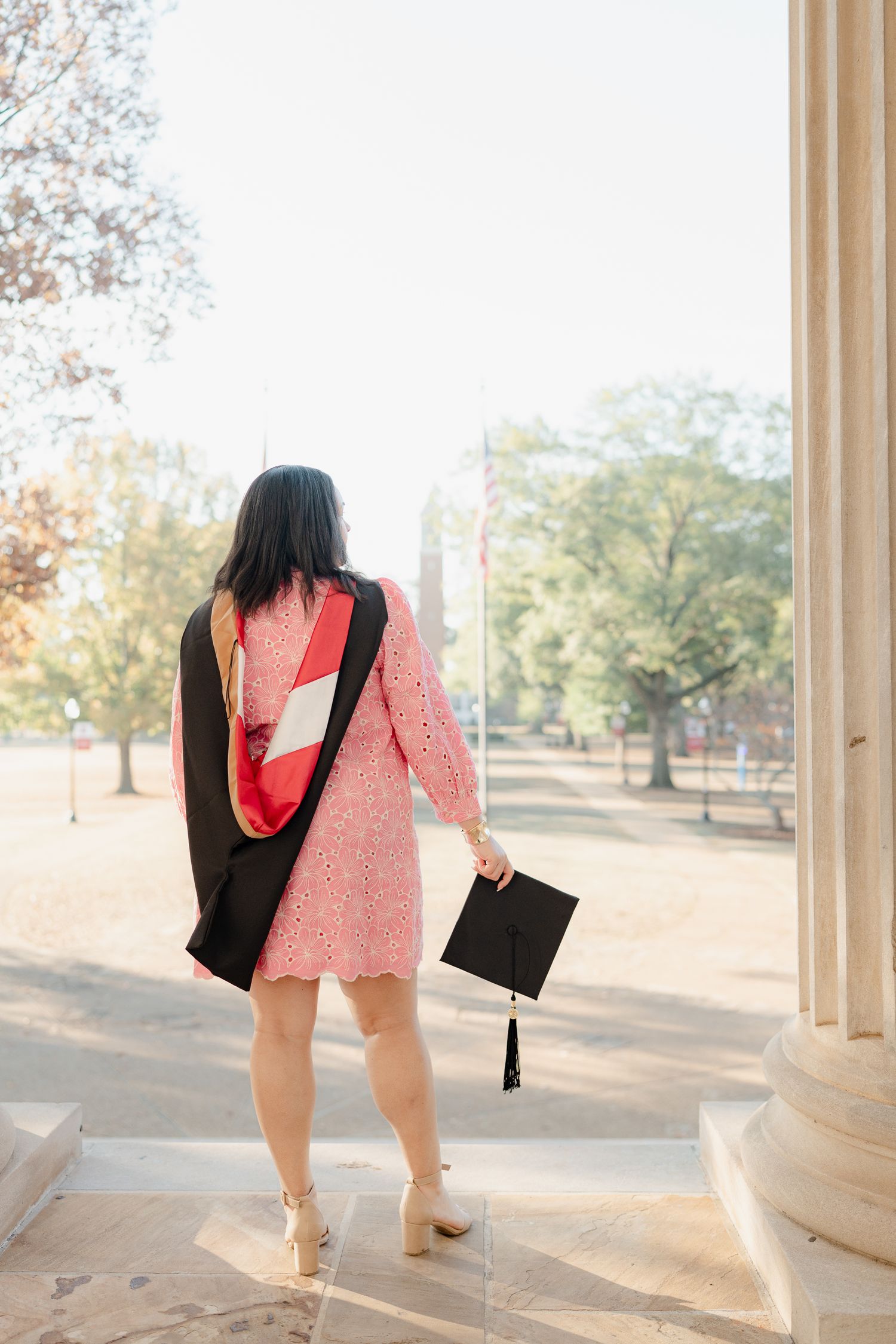 A graduate in academic regalia and pink dress poses between white columns on campus.