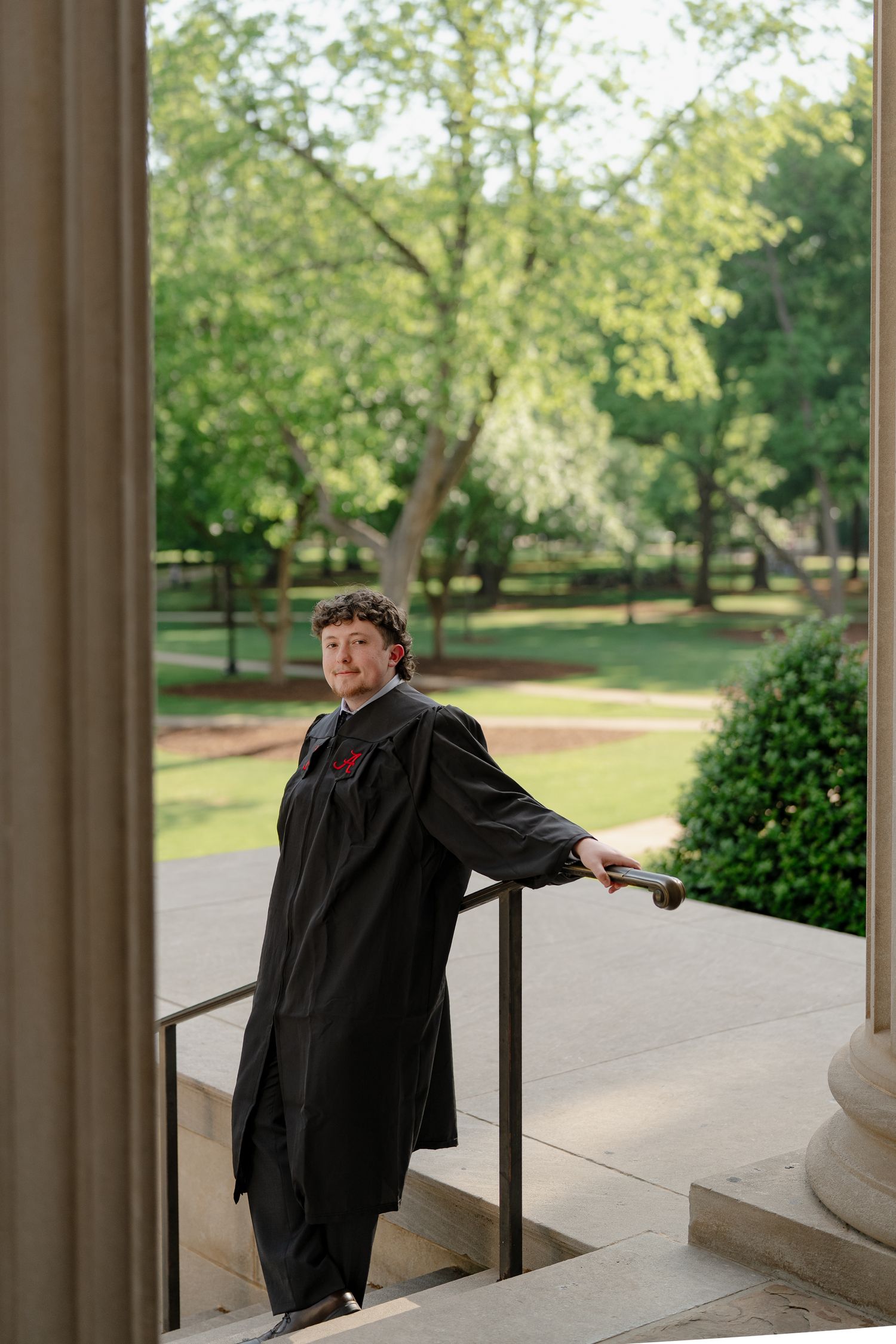 Graduate in black gown poses on stone steps between columns with lush green campus trees in background.