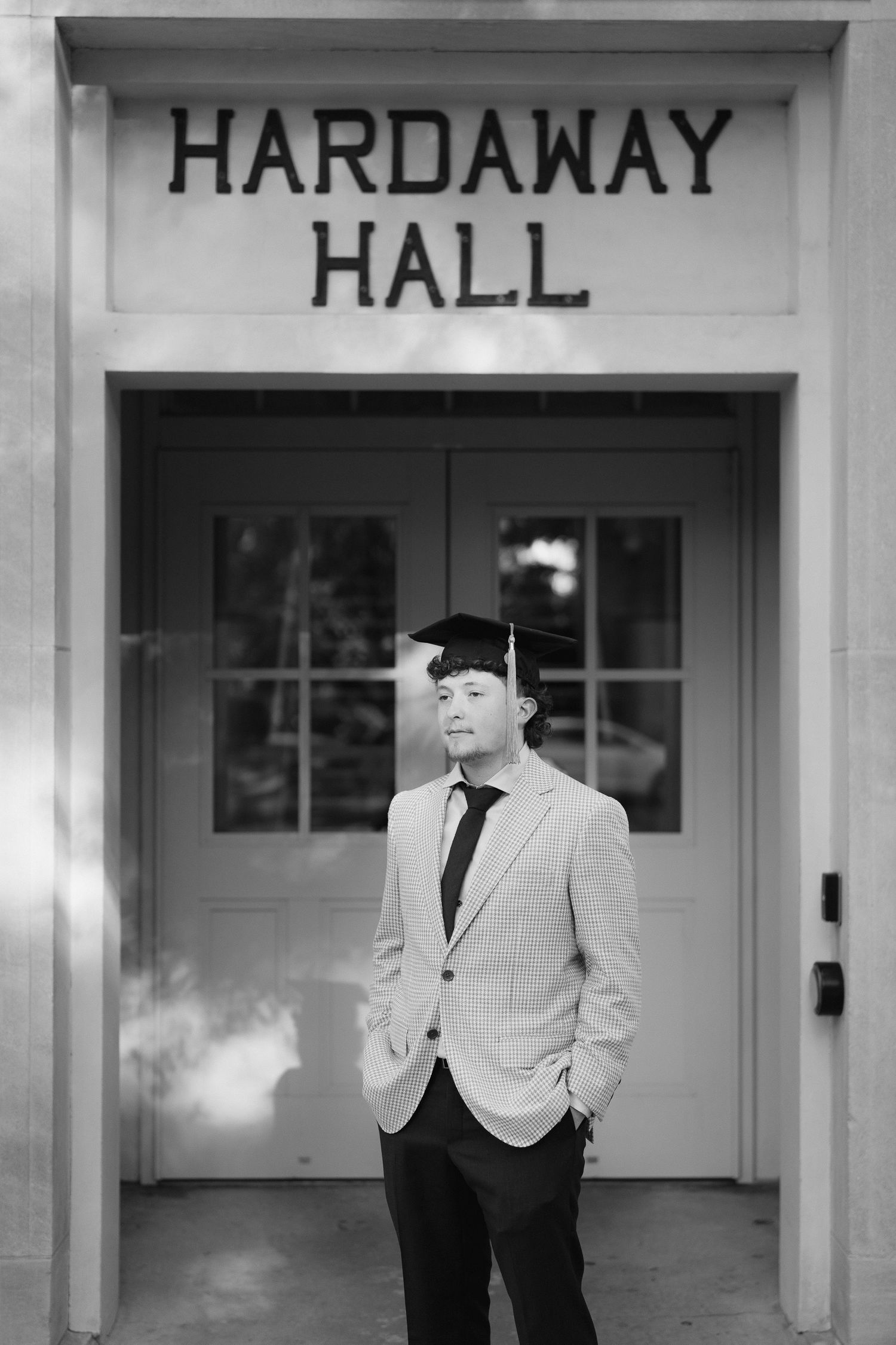 Graduate posing in cap and blazer under Hardaway Hall sign, black and white photo.