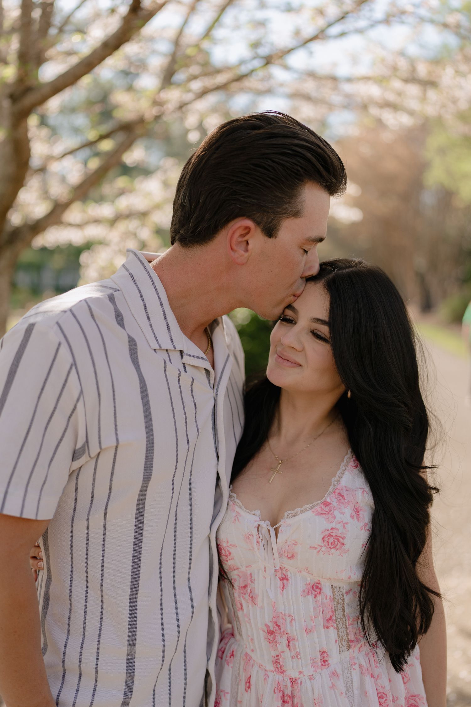 Man kissing woman on forehead as she smiles softly under blooming spring cherry blossom trees.