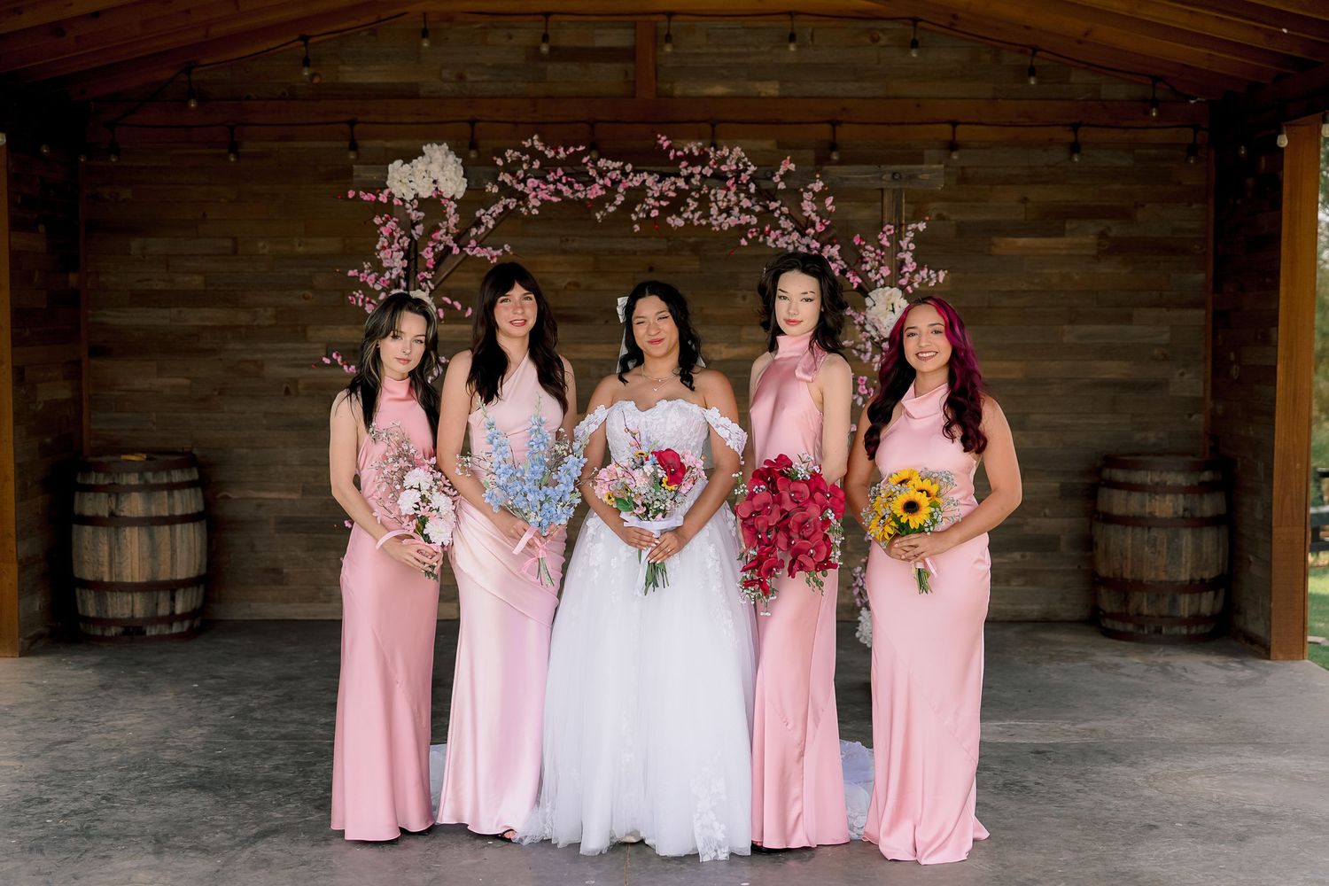 Bride in white gown poses with four bridesmaids in pink dresses under floral arch in rustic barn.