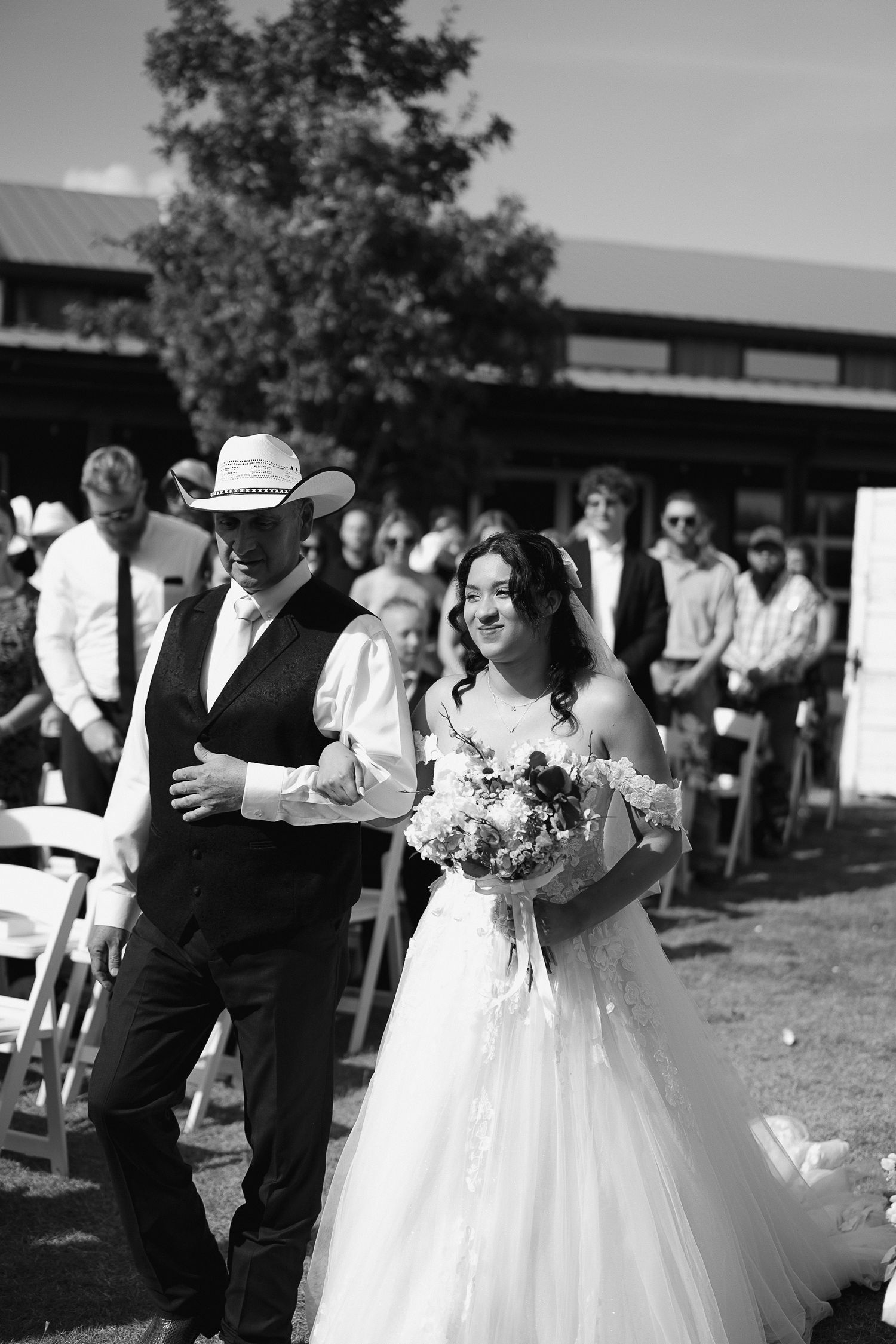 Bride walks down outdoor aisle in wedding gown holding bouquet, escorted by man in cowboy hat and vest.