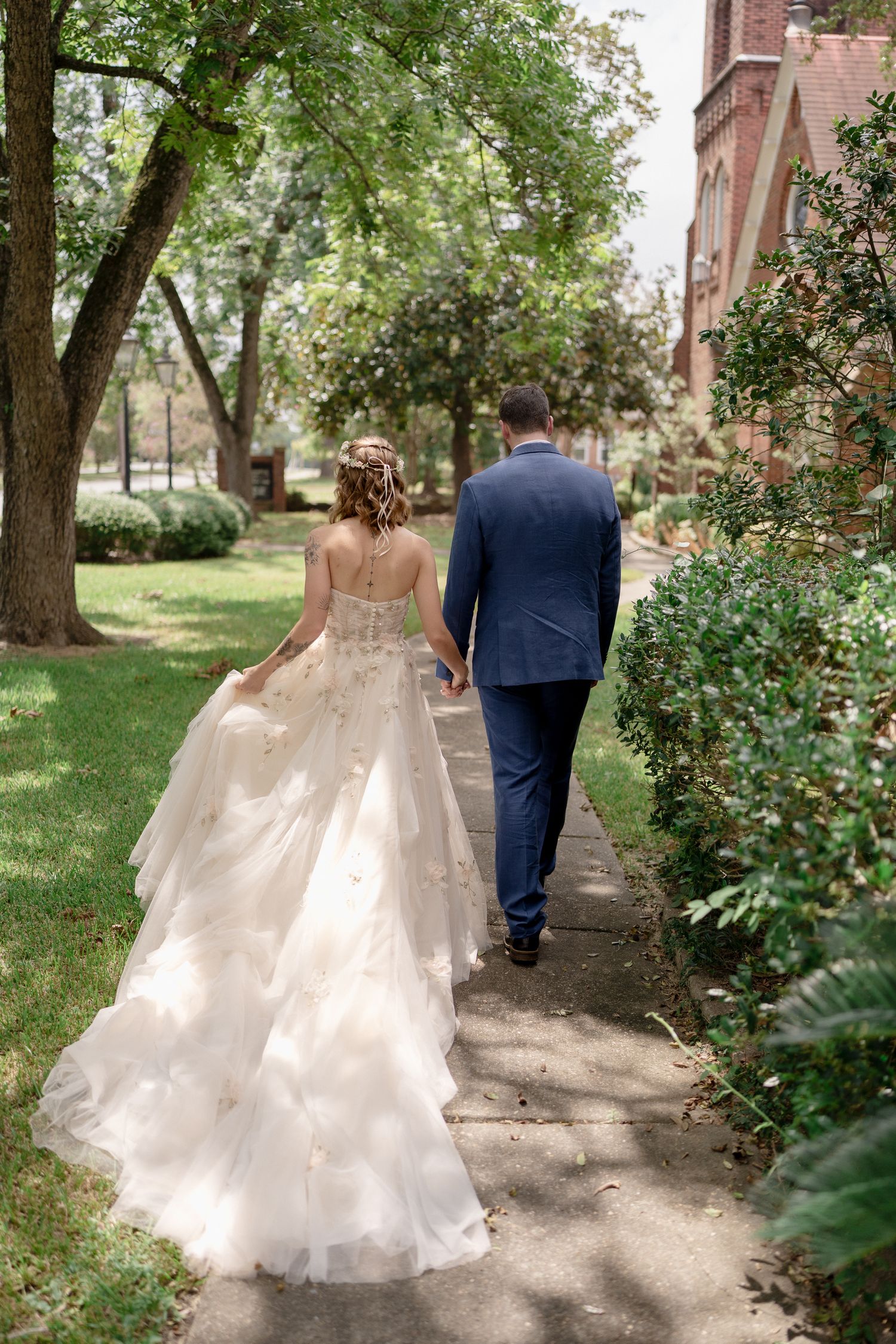 Bride and groom walking hand in hand down a garden path, bride in layered ballgown, groom in navy suit.