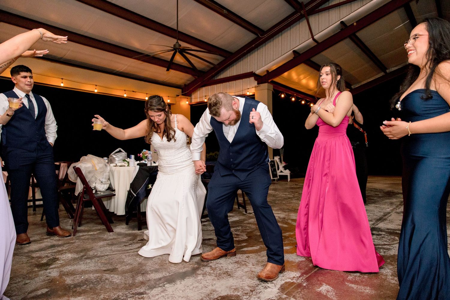 Bride and groom dance energetically with wedding guests in pink and navy attire at indoor reception.