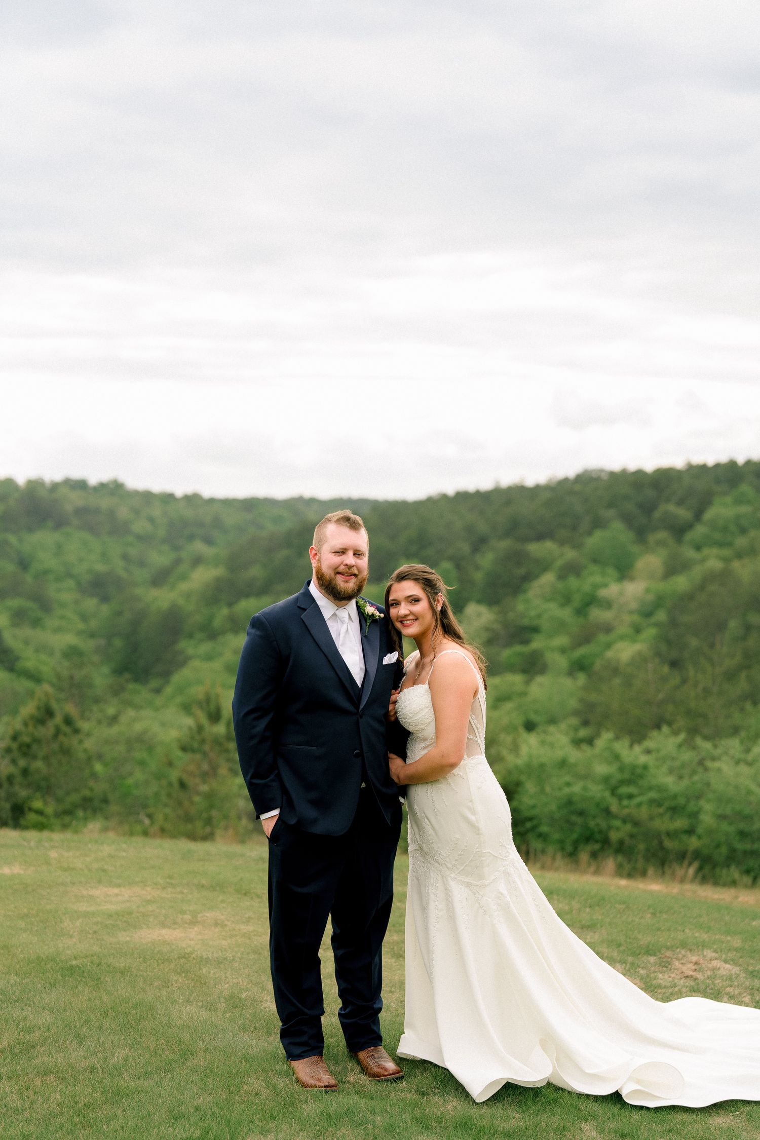 Bride and groom pose together on a grassy hill with lush green forest in the background.