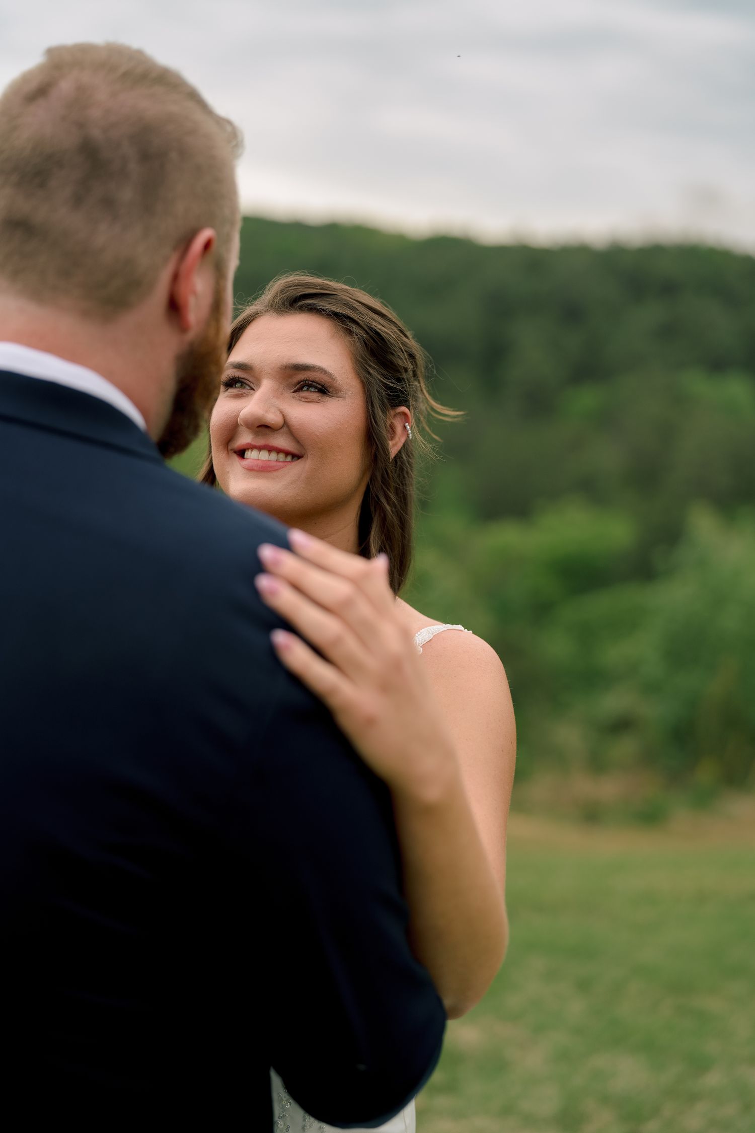 Smiling bride in white dress gazes lovingly at groom in navy suit outdoors with green hills in background.