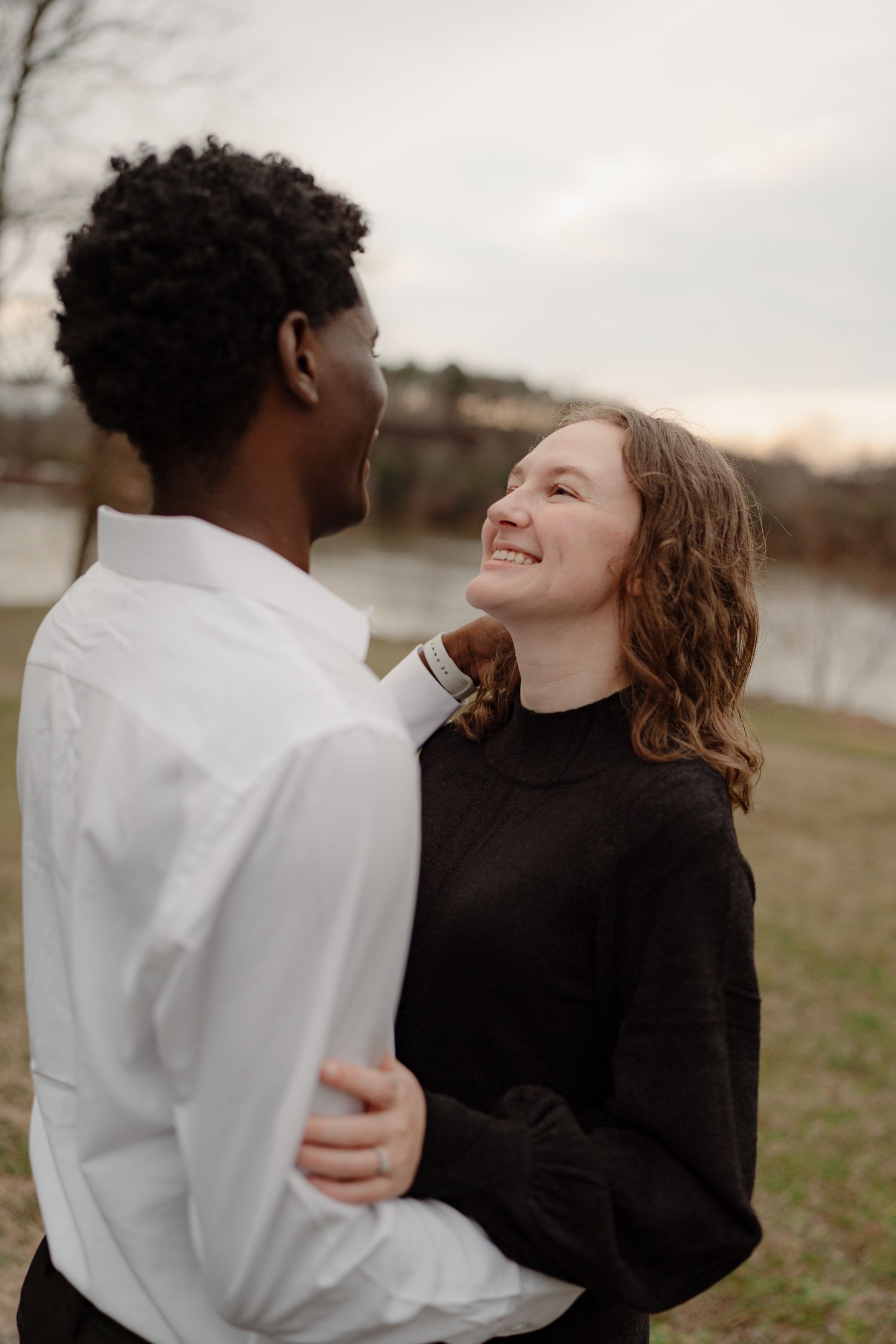 A woman in a black sweater smiles warmly at her partner near a river during a romantic outdoor photo session.