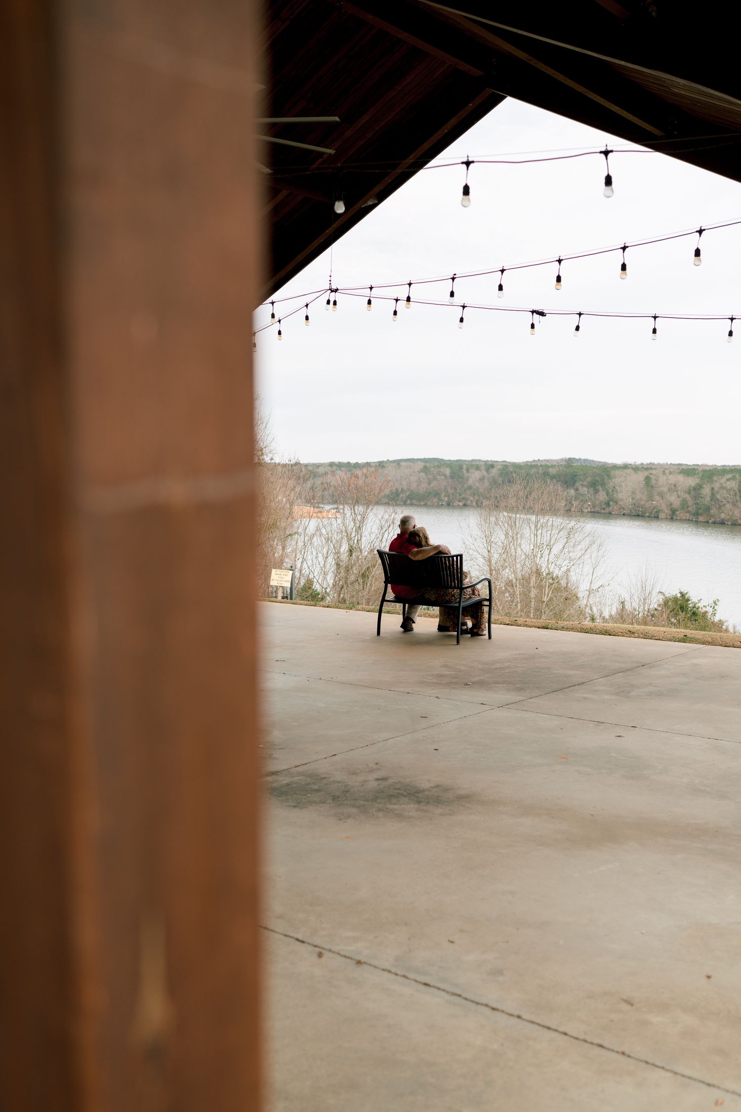 Couple sitting together in chairs on a patio overlooking a scenic lake view with string lights above.