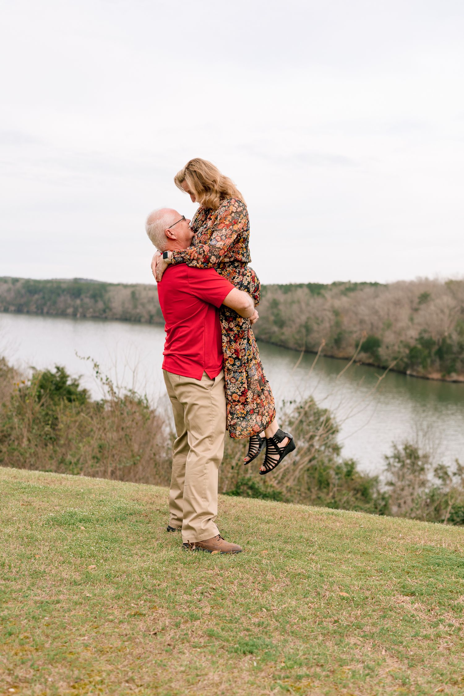 Man in red shirt joyfully lifting woman in floral dress near a scenic lake overlook.