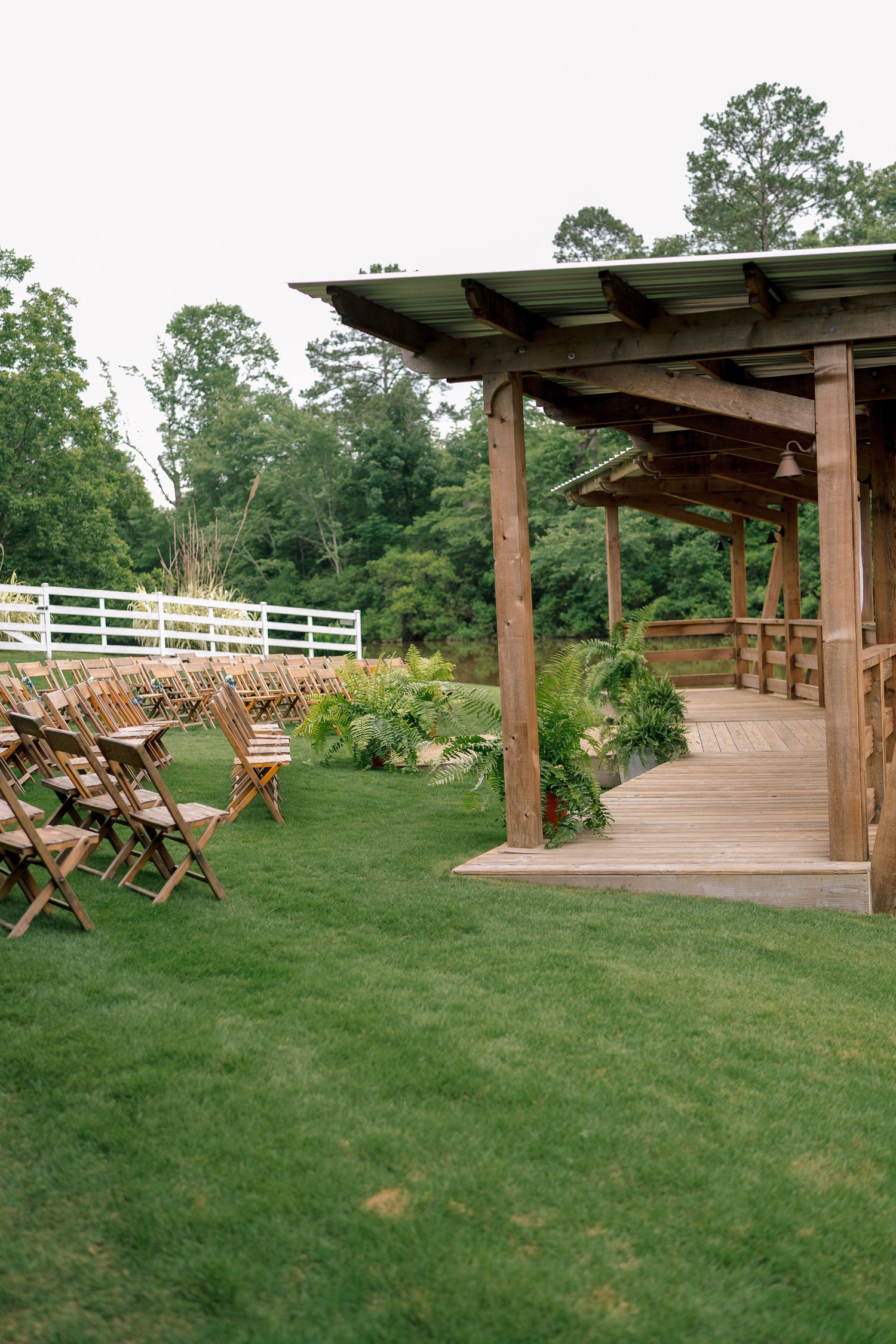Rustic outdoor wedding ceremony setup with wooden folding chairs and wooden pavilion surrounded by lush greenery.