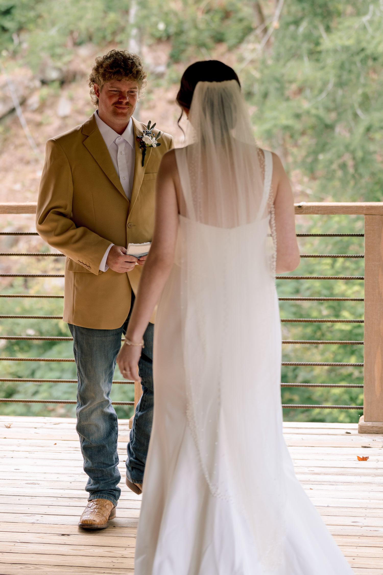 Groom in tan blazer and jeans walks toward bride in white wedding gown on wooden deck during first look.
