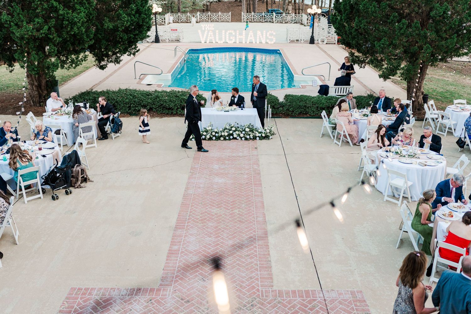 Aerial view of an outdoor wedding reception with pool and white tables near a grand building.