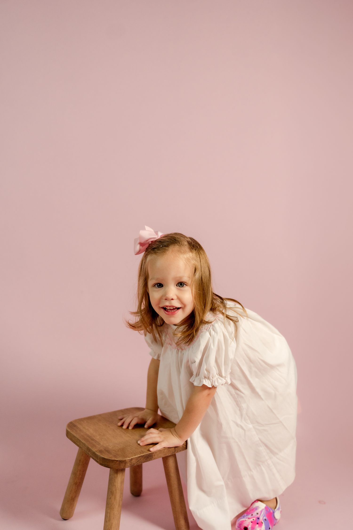 A playful moment captured as a small child interacts with a wooden stool against pink.
