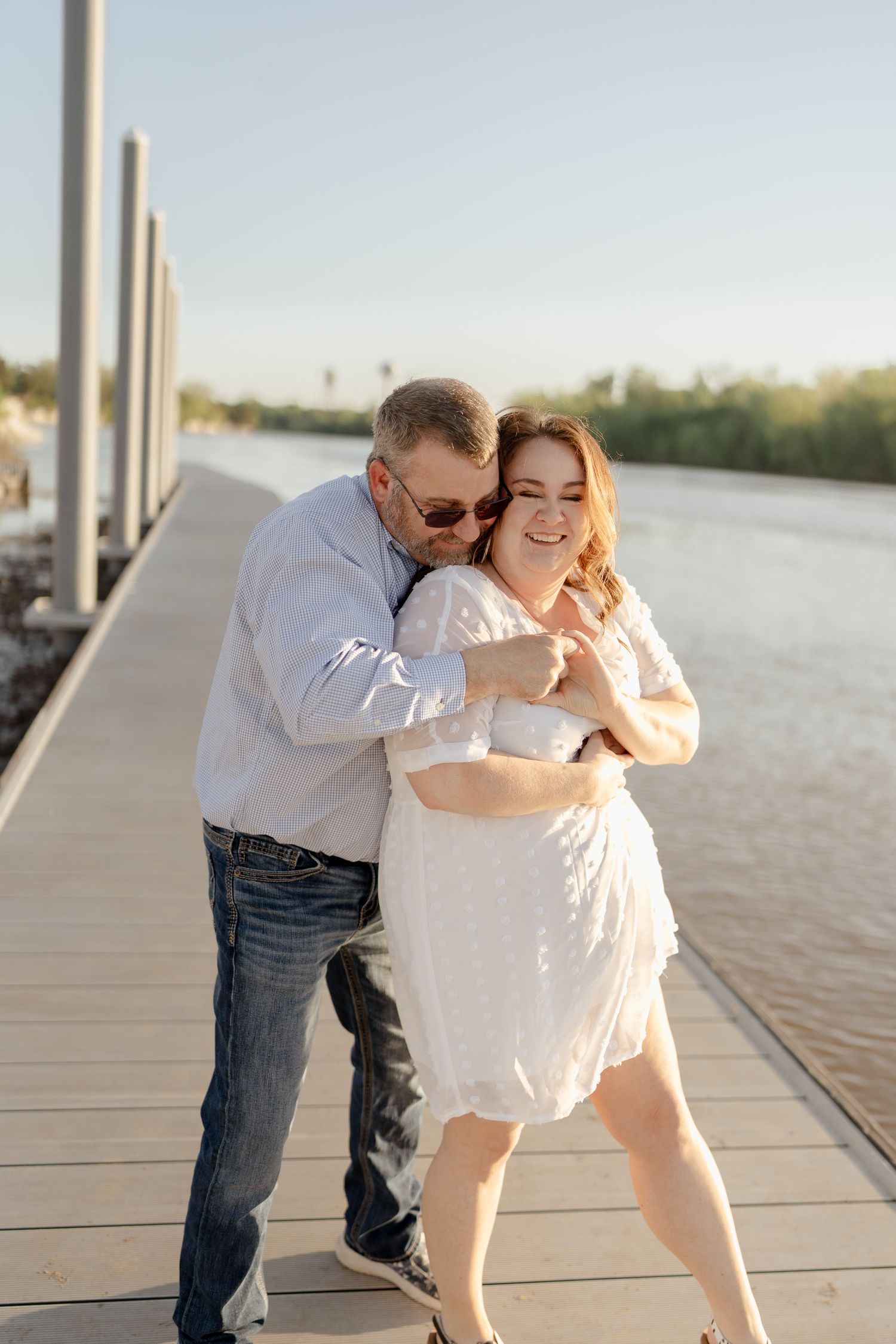 Two people share a romantic embrace while laughing on a waterfront deck.
