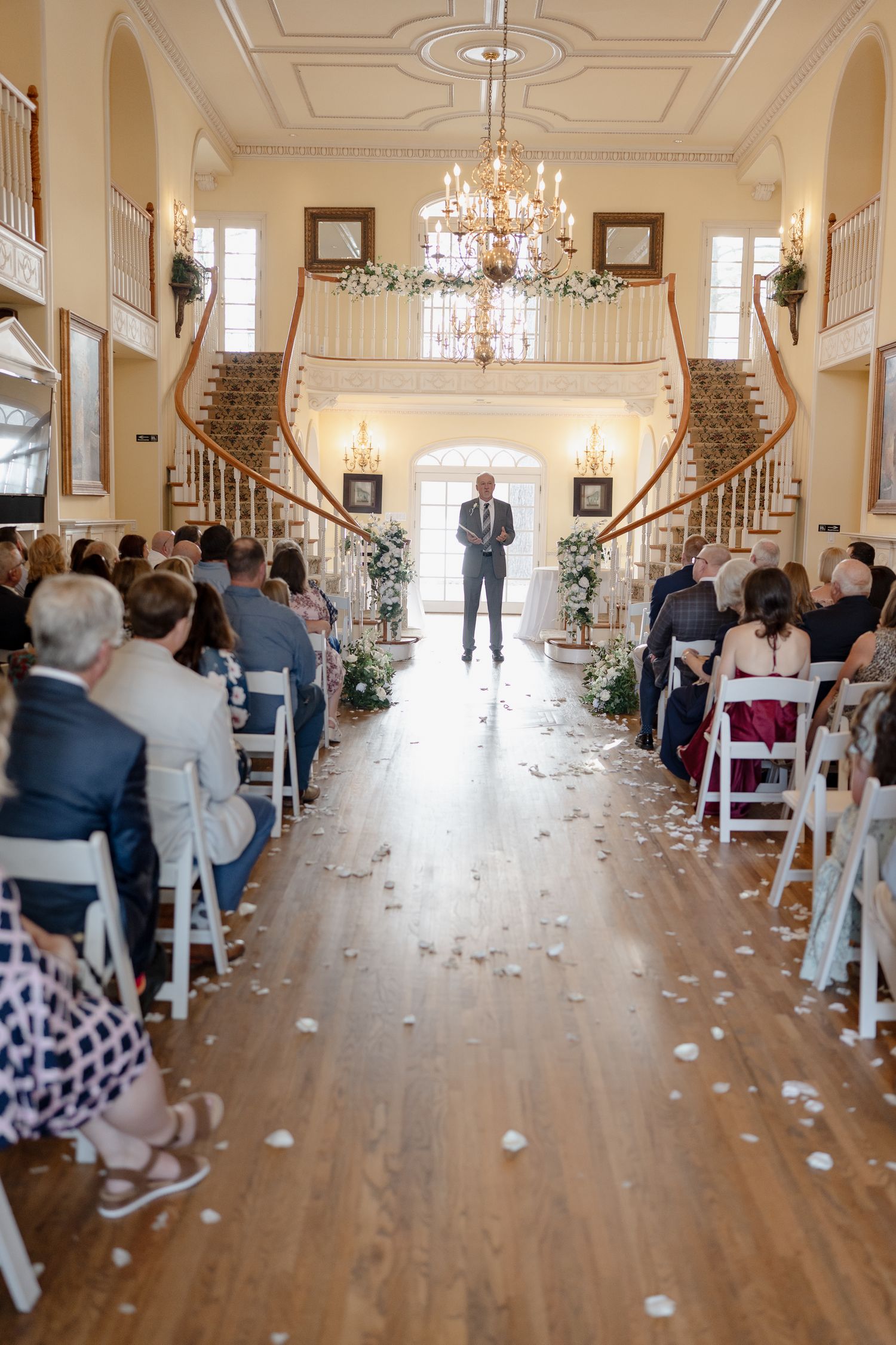 A wedding ceremony setup in an elegant ballroom with white chairs, flower petals, and crystal chandeliers.
