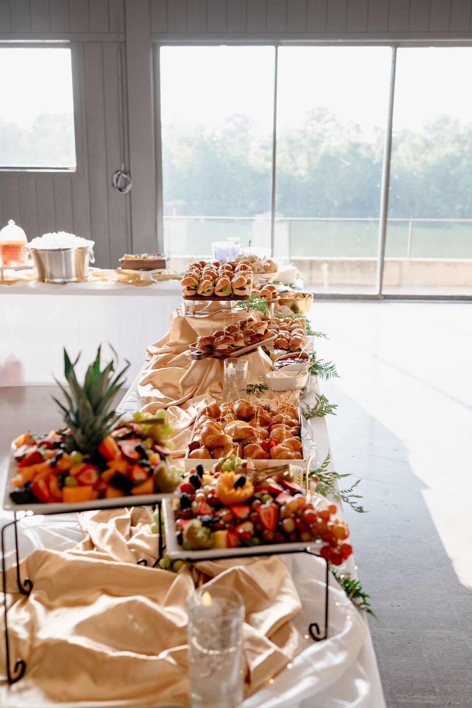 Elegant catering display with food platters arranged on white tablecloths near large windows.