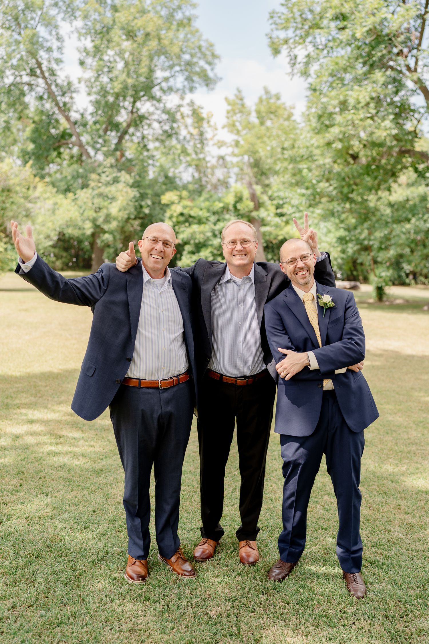Three people in navy suits stand together smiling outdoors on grass with green trees in the background.
