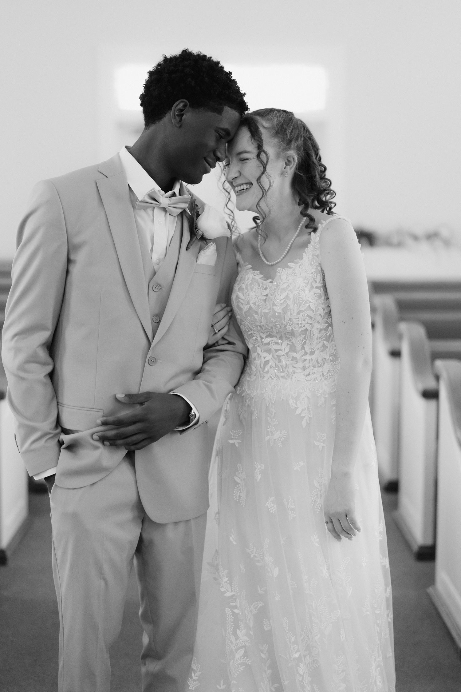Black and white photo of interracial bride and groom laughing together in church aisle.