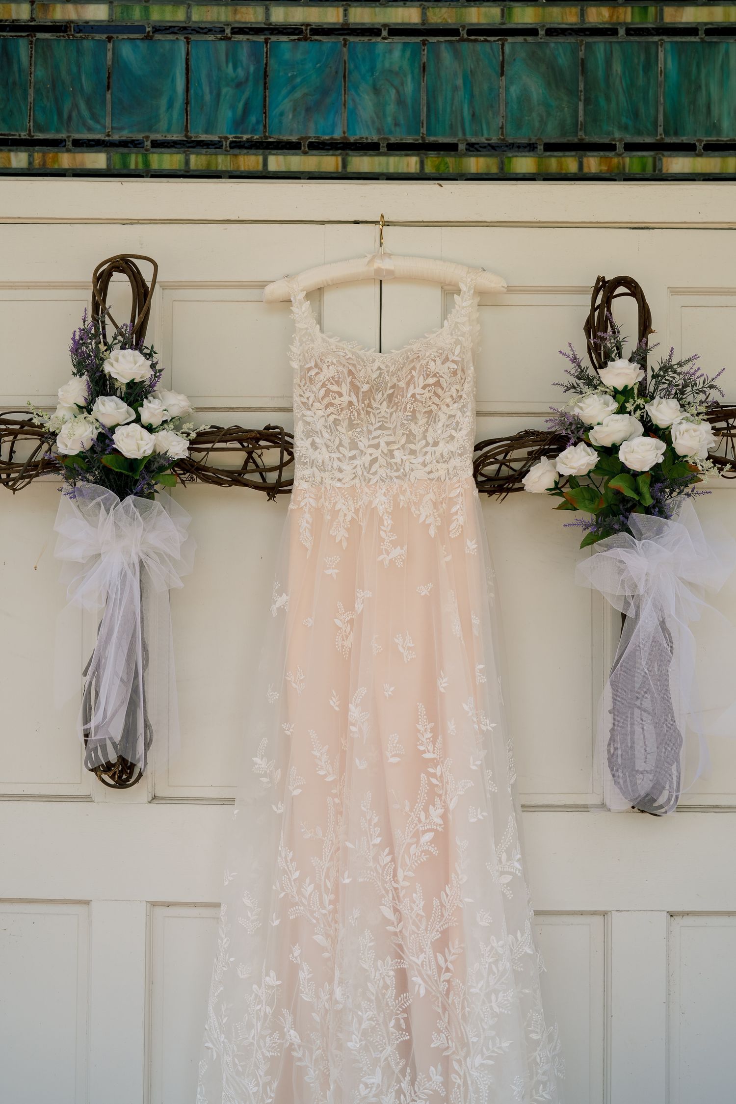 Blush lace wedding dress hanging on church door flanked by rustic twig wreaths with white floral bouquets and tulle bows.