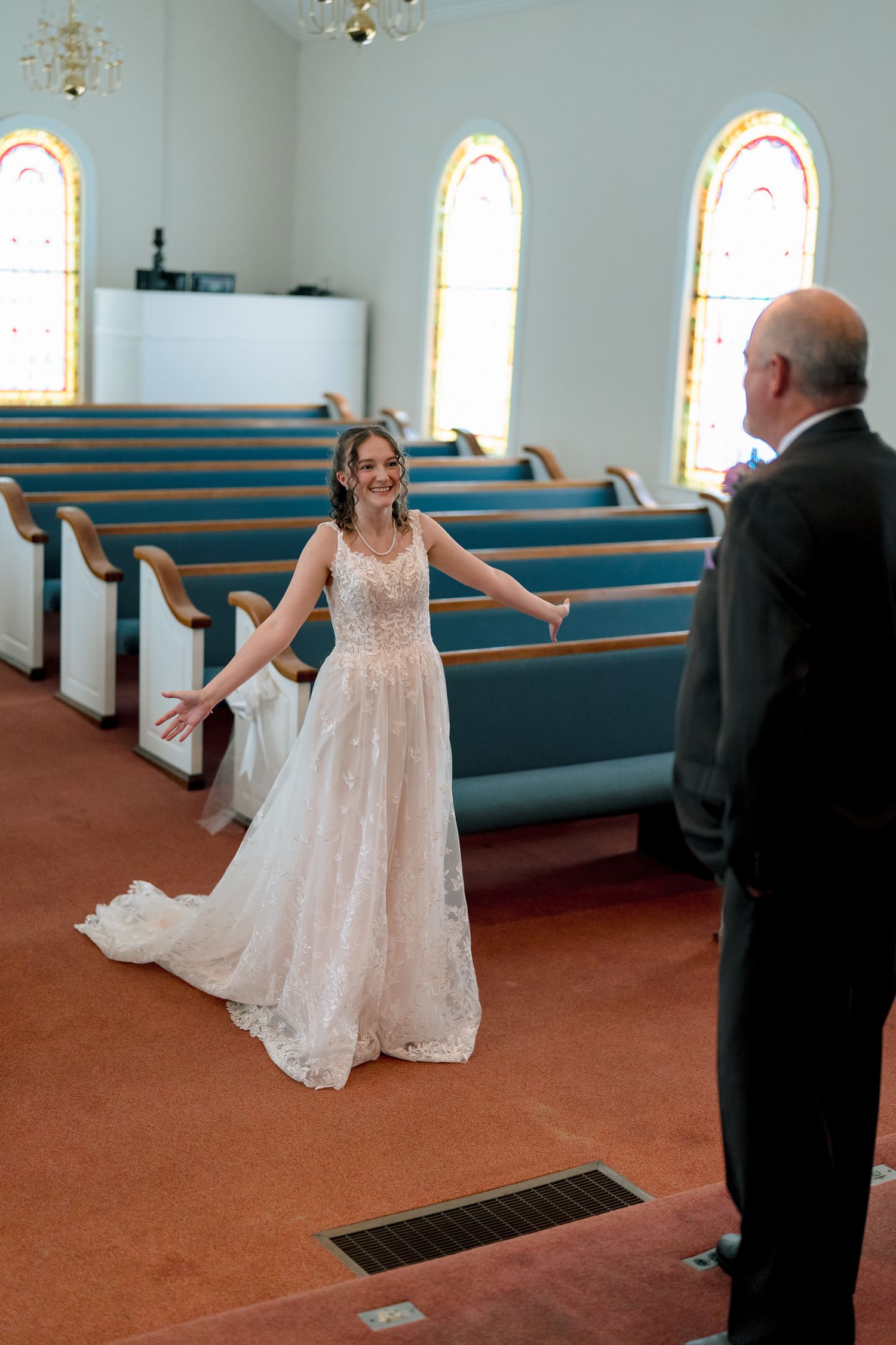 Bride in lace wedding gown spreading arms wide with joy in a church aisle with blue pews and stained glass windows.