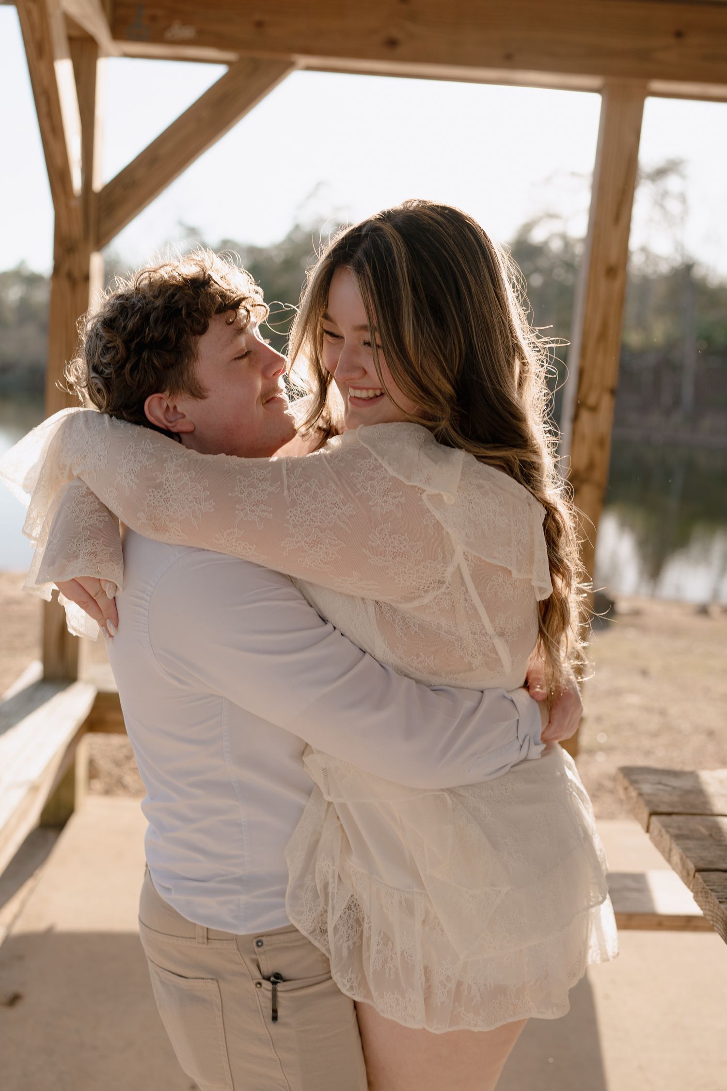Couple embracing and smiling at each other under a wooden pergola with a lake visible in the background.
