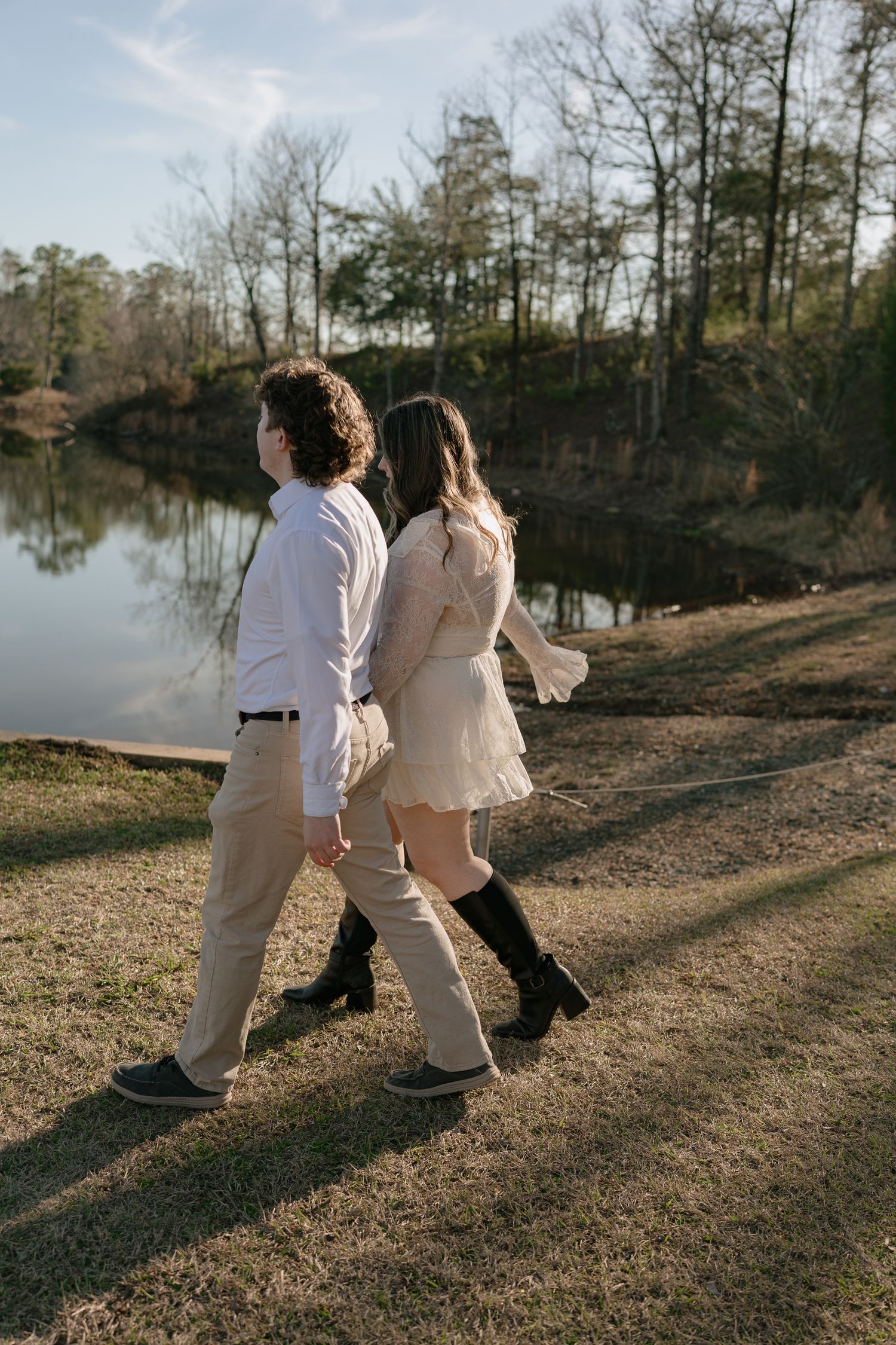 Engaged couple walking hand in hand along a serene lakeside path in golden hour light.