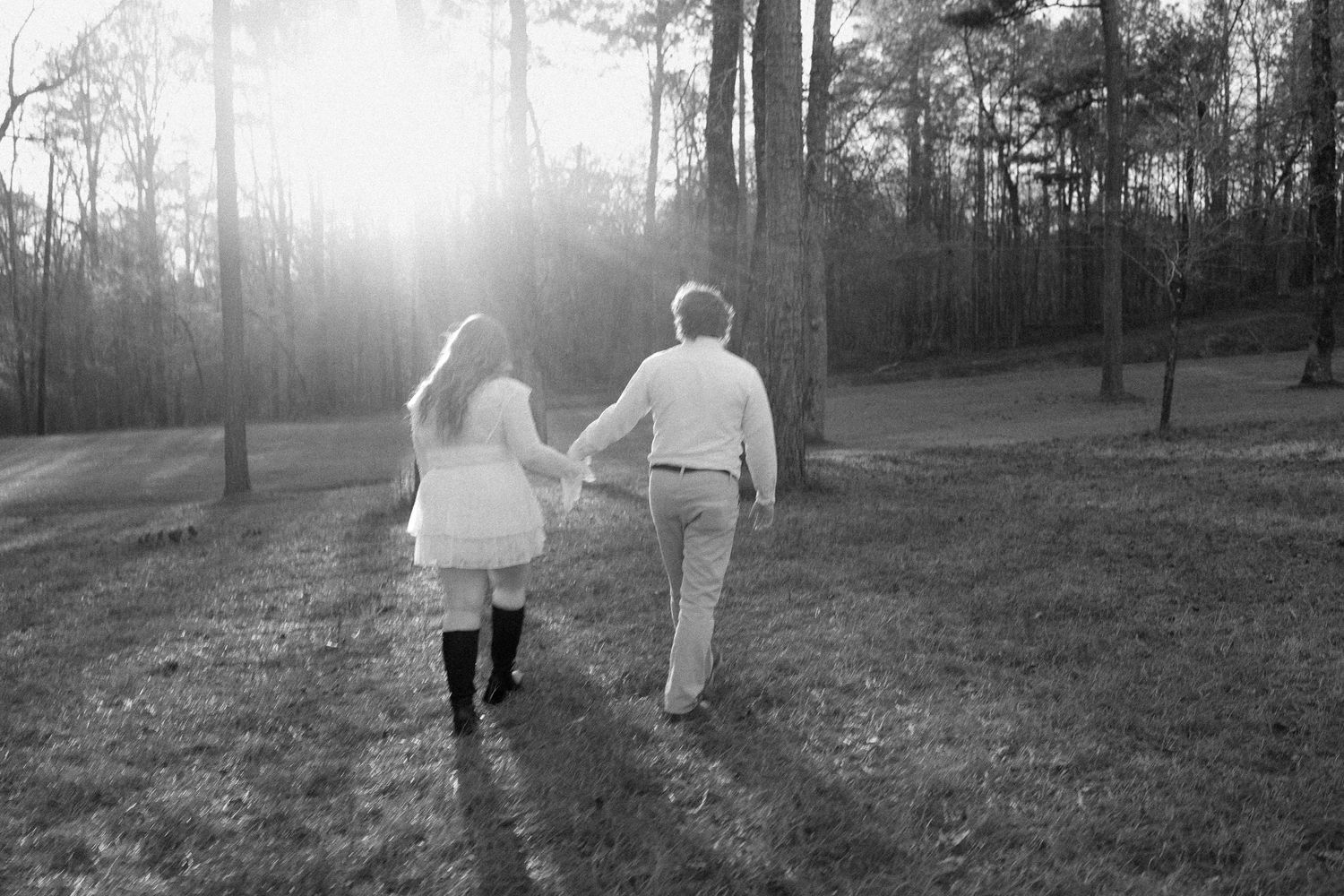 Black and white photo of couple walking hand in hand through a glowing sun-drenched forest path.
