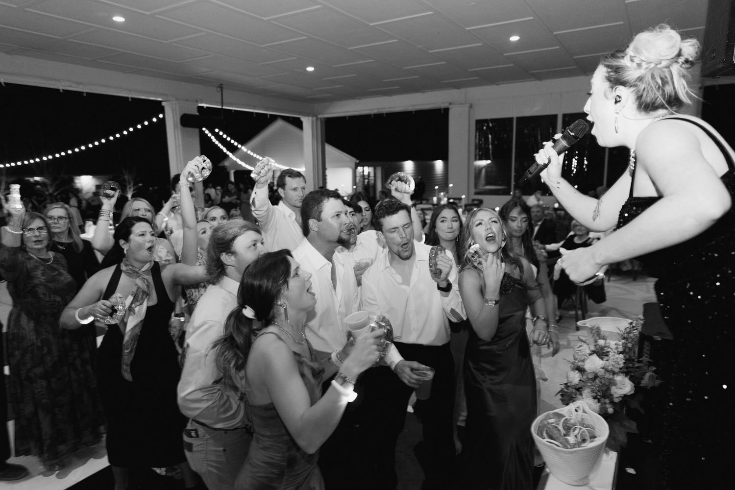 Black and white photo of wedding guests cheering as bride tosses bouquet at elegant indoor reception.