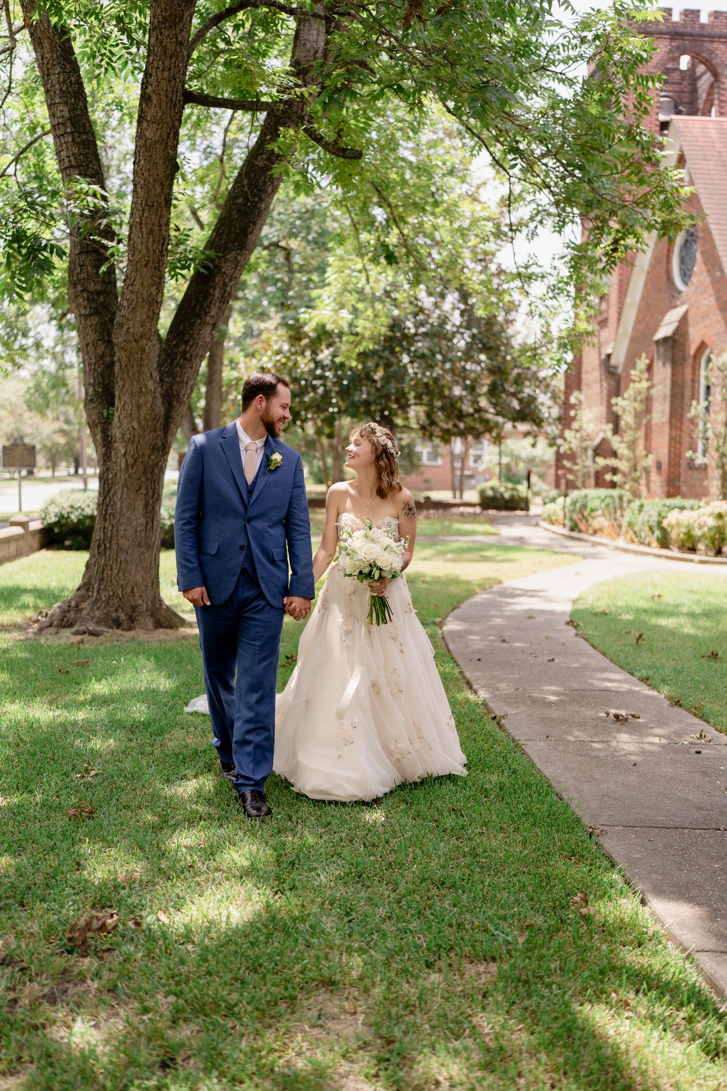 A couple walks together on a pathway near a brick church on a sunny summer day, surrounded by lush trees.