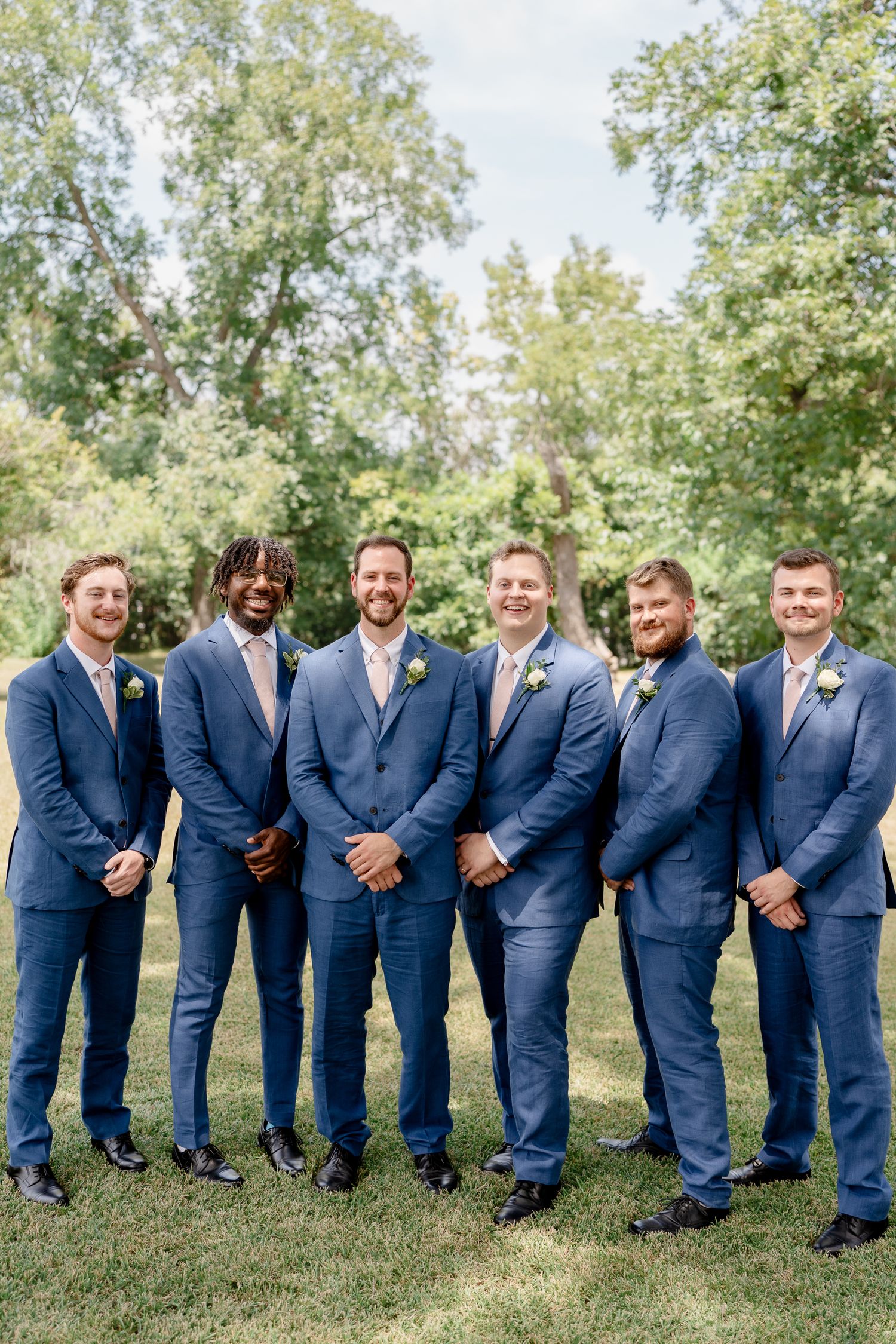 A wedding party in matching navy blue suits poses in a creative curved line formation on green grass outdoors.