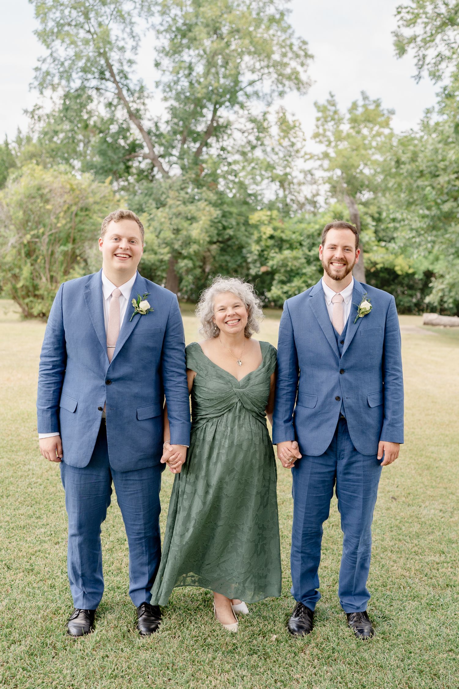 Three people in formal attire stand together outdoors, with two wearing navy suits and one in a green dress.
