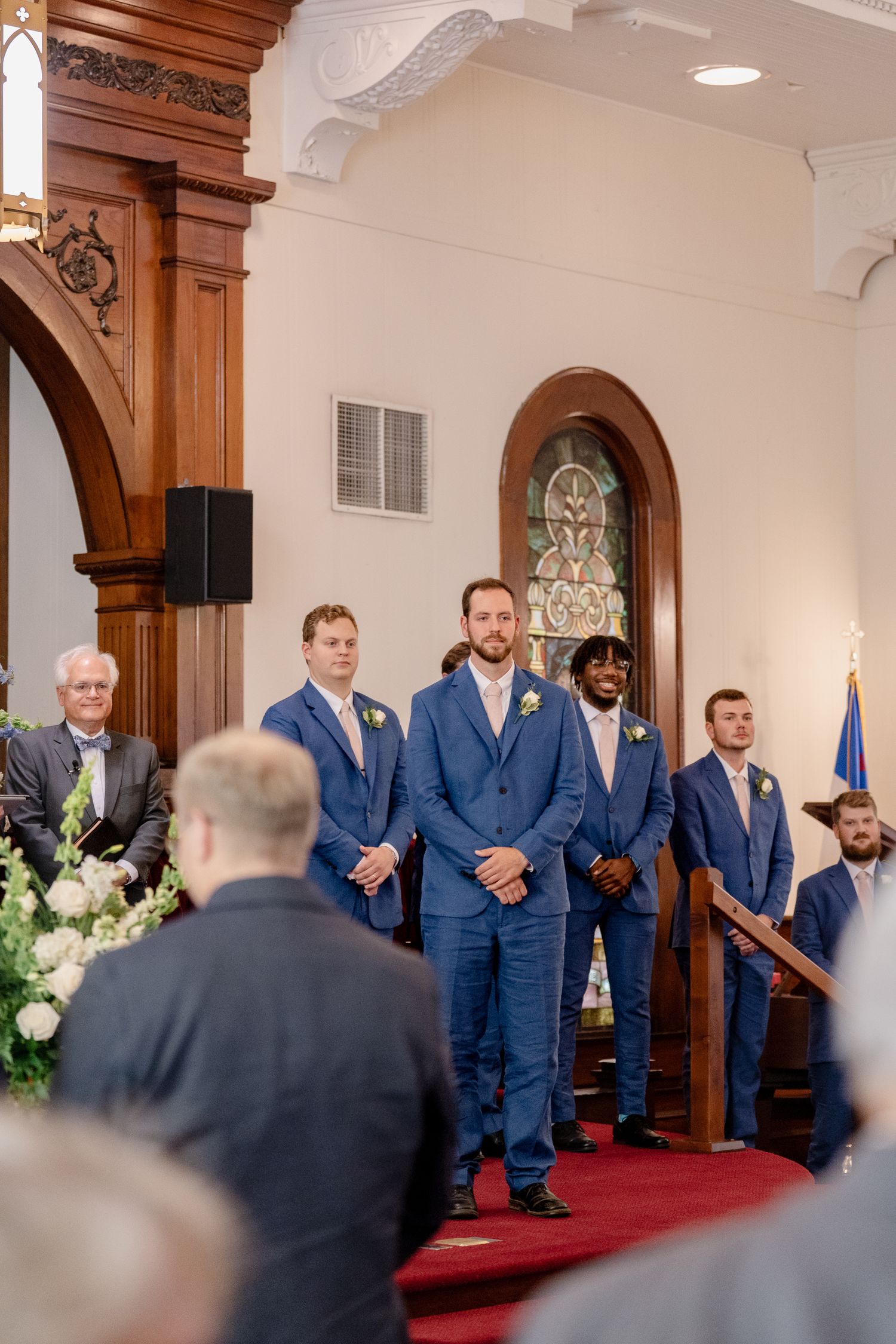 Wedding party stands at altar in church with wooden archways and stained glass window during ceremony.