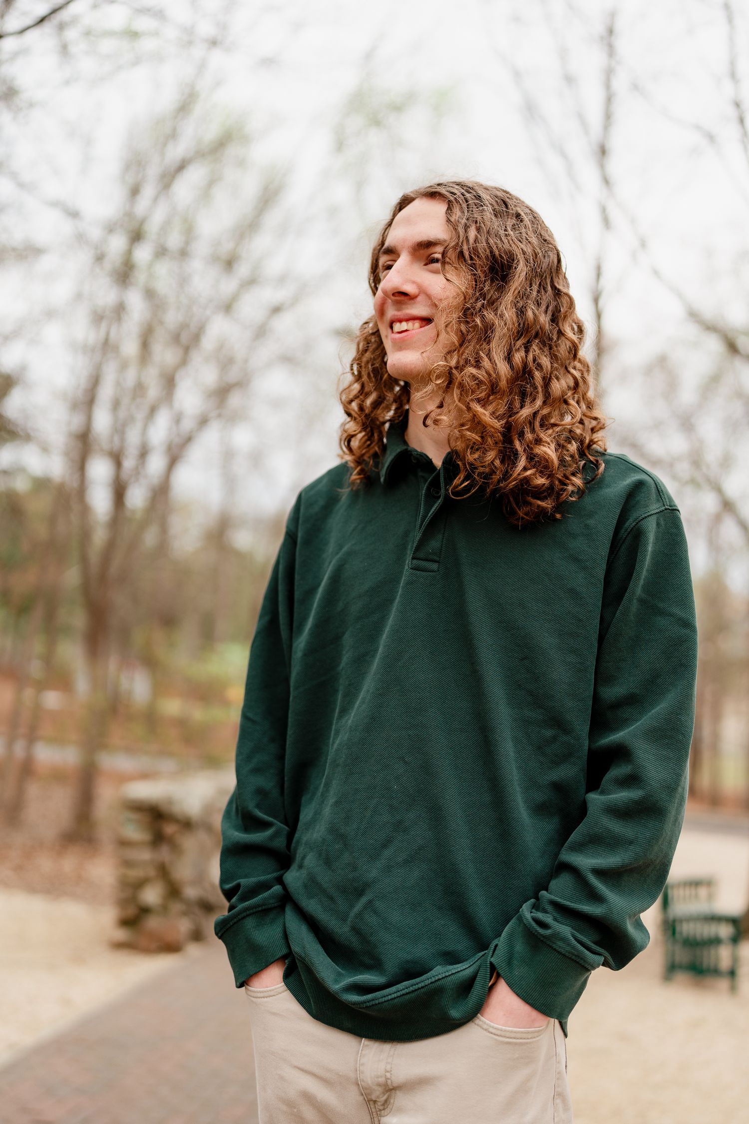 Person wearing dark green sweater standing outdoors on brick pathway with trees in background.