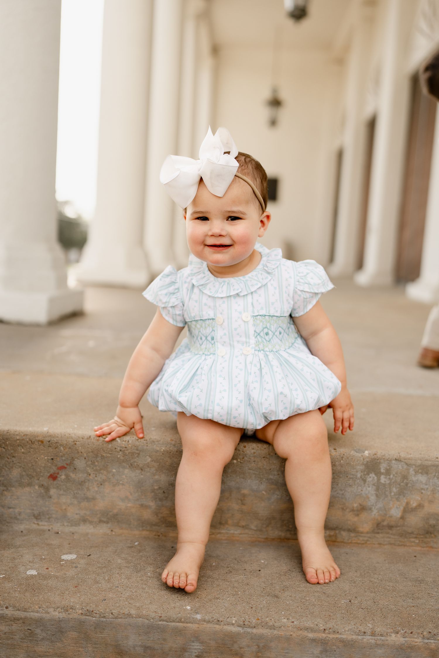 Baby wearing a light blue dress and white bow sits smiling on concrete steps.