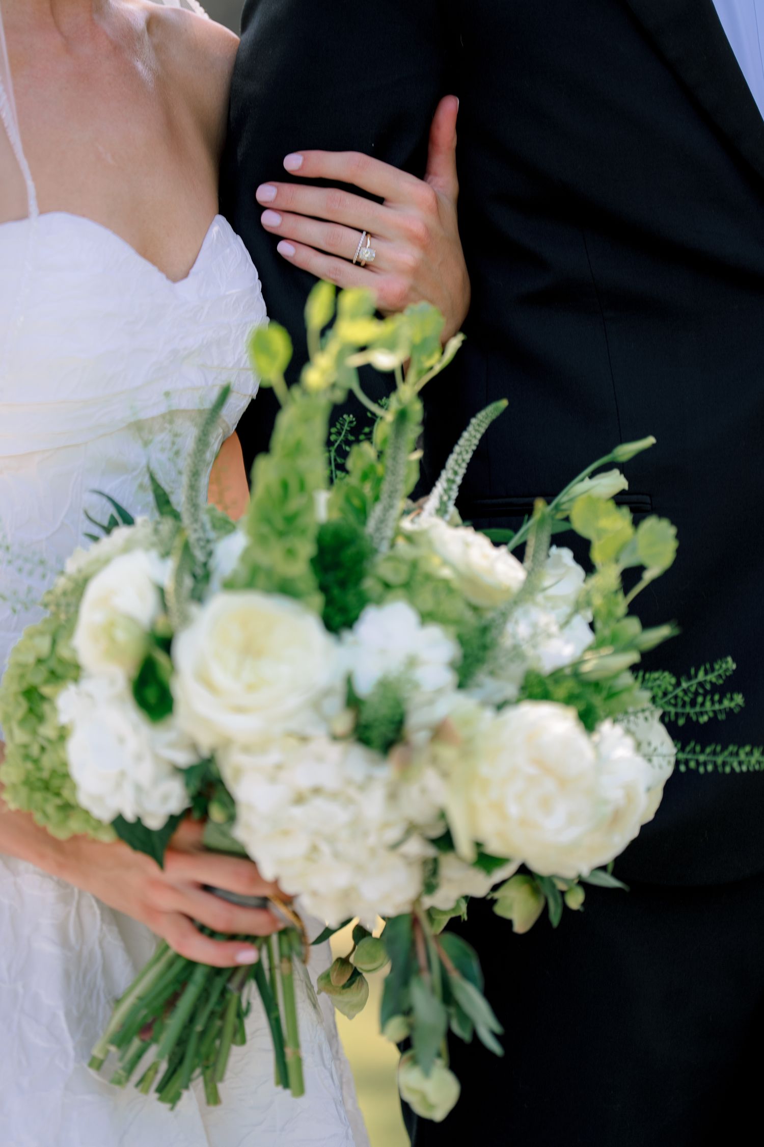 Bride holding white and green wedding bouquet with roses while groom in black suit stands beside her.