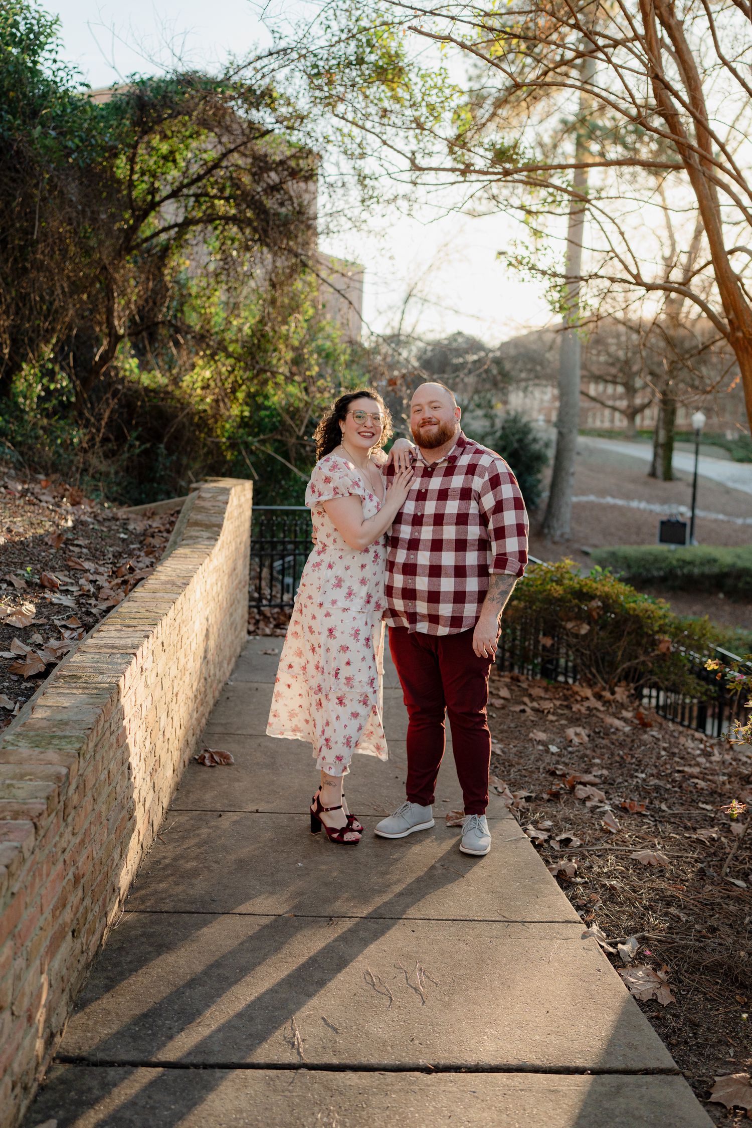 Couple poses together on stone-walled pathway during golden hour in an urban park setting.