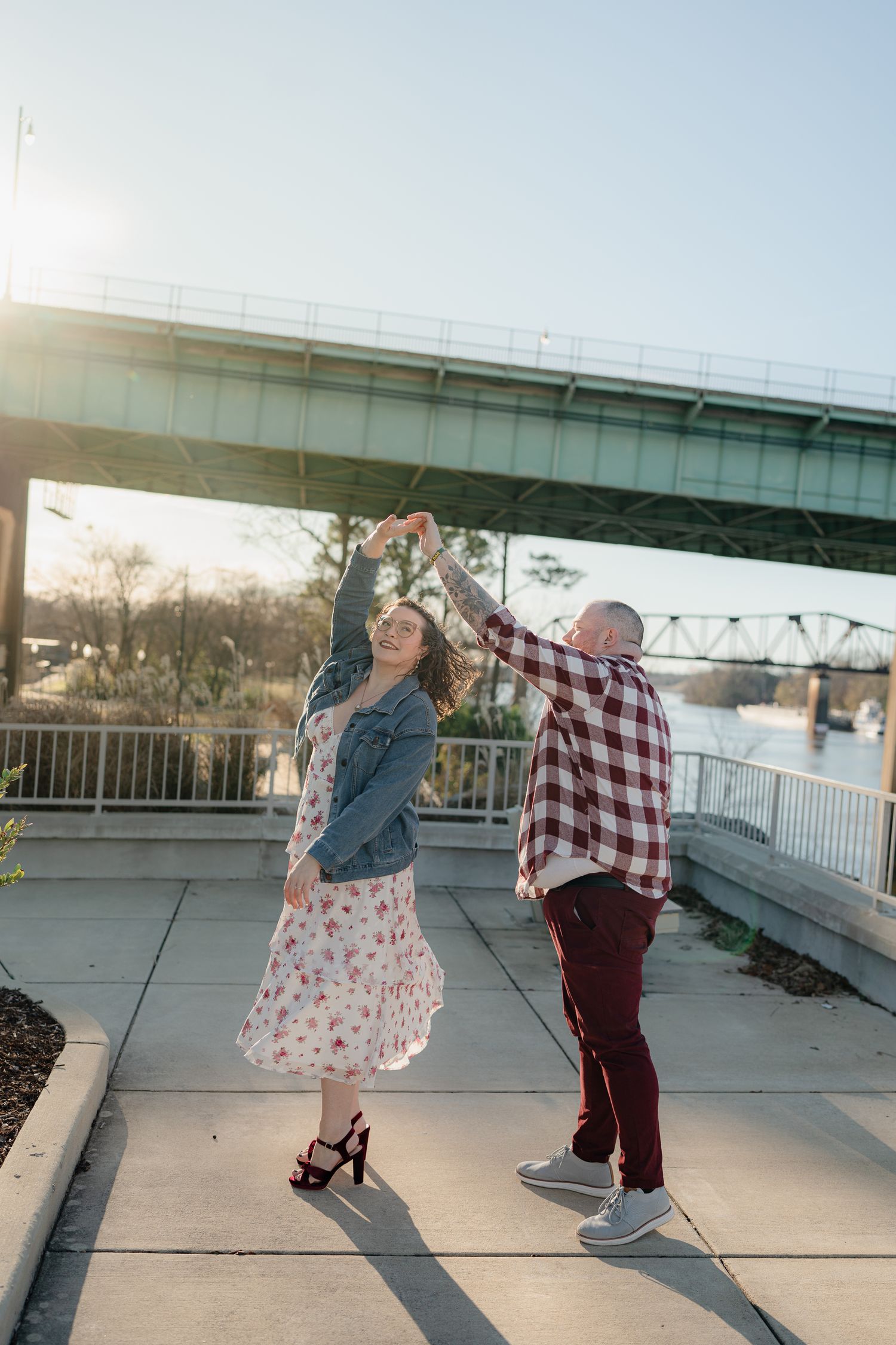 A couple dances together on a concrete patio with a bridge visible in the background during sunset.