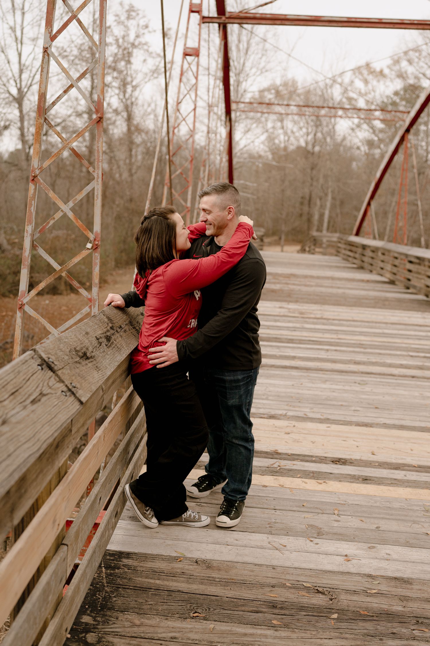 A couple embraces on a wooden bridge surrounded by bare trees in muted tones.
